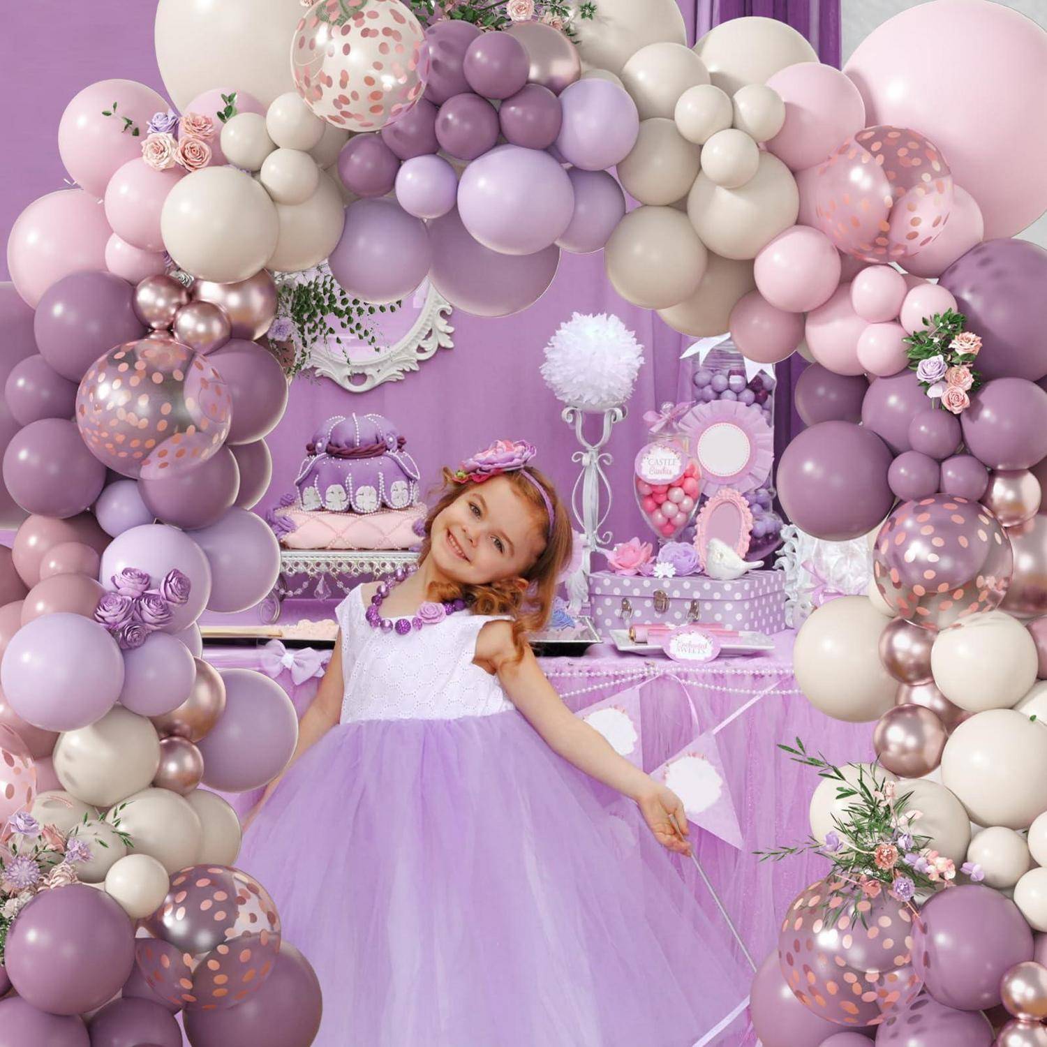 Junges Mädchen in einem lavendelfarbenen Kleid lächelt bei einer lila Geburtstagsfeier und steht unter einem Pastellballonbogen, während es einen Zauberstab hält.
