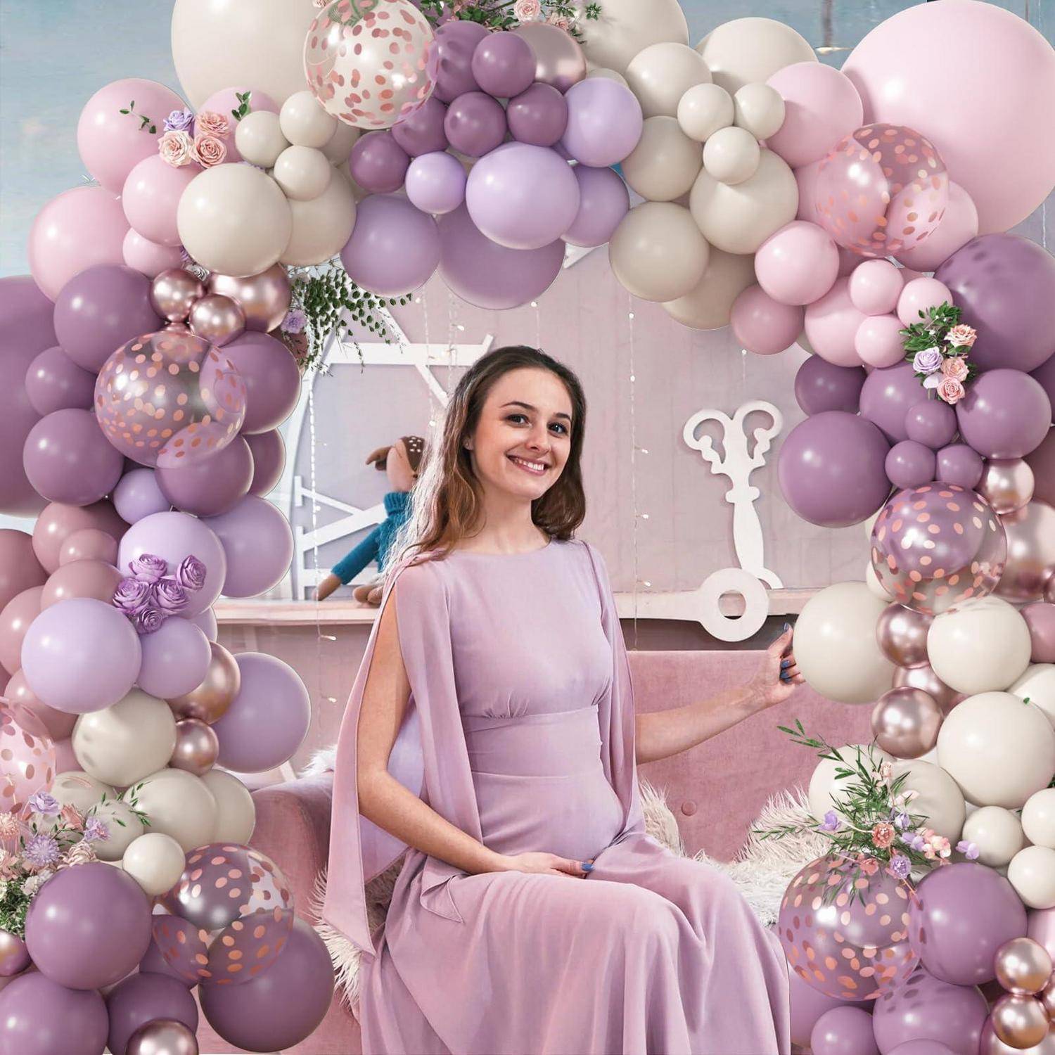 Lächelnde Frau in einem Lavendelkleid sitzt unter einem pastellvioletten und cremefarbenen Ballonbogen vor einer dekorierten Partykulisse.