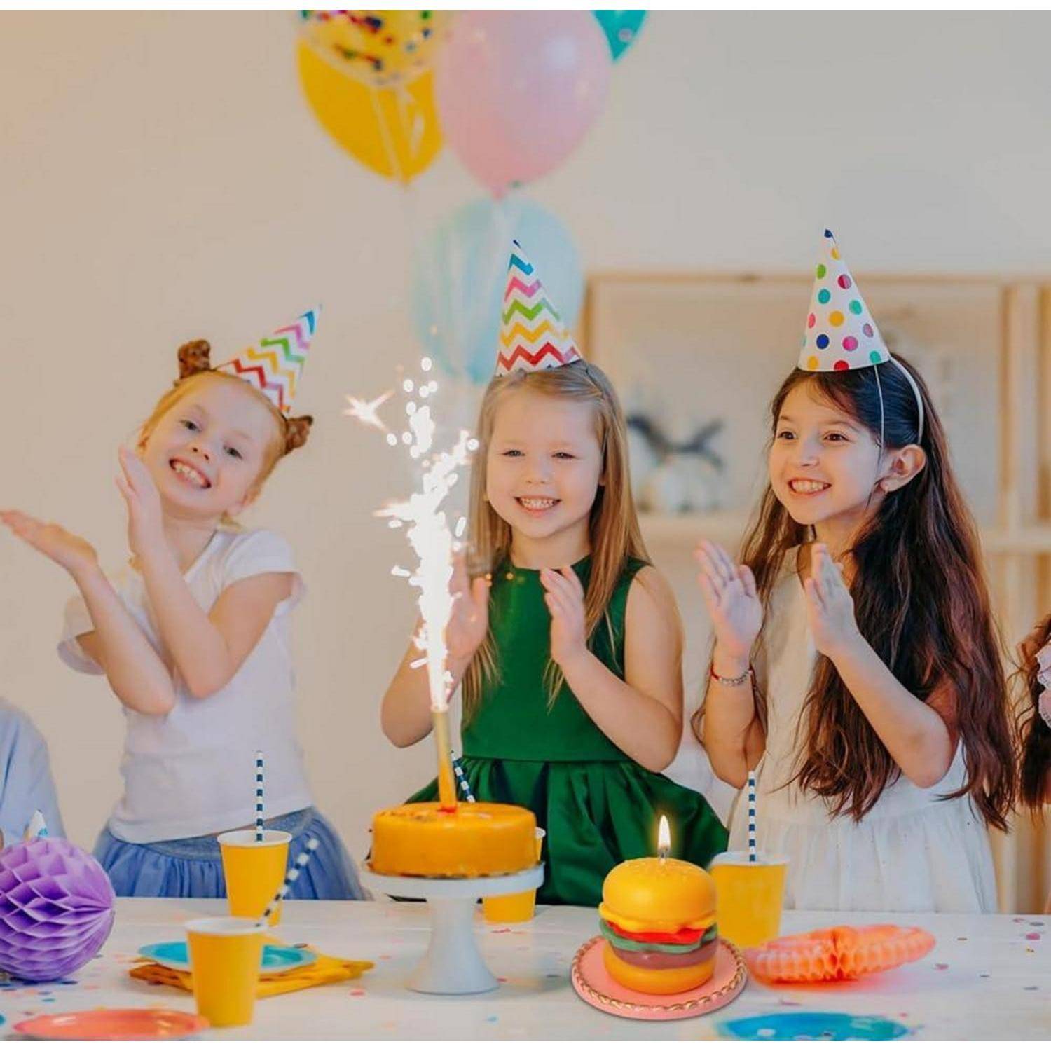 Drei Mädchen mit Partyhüten klatschen neben einer Geburtstagstorte mit einem Wunderkerzen und bunten Partydekoration.