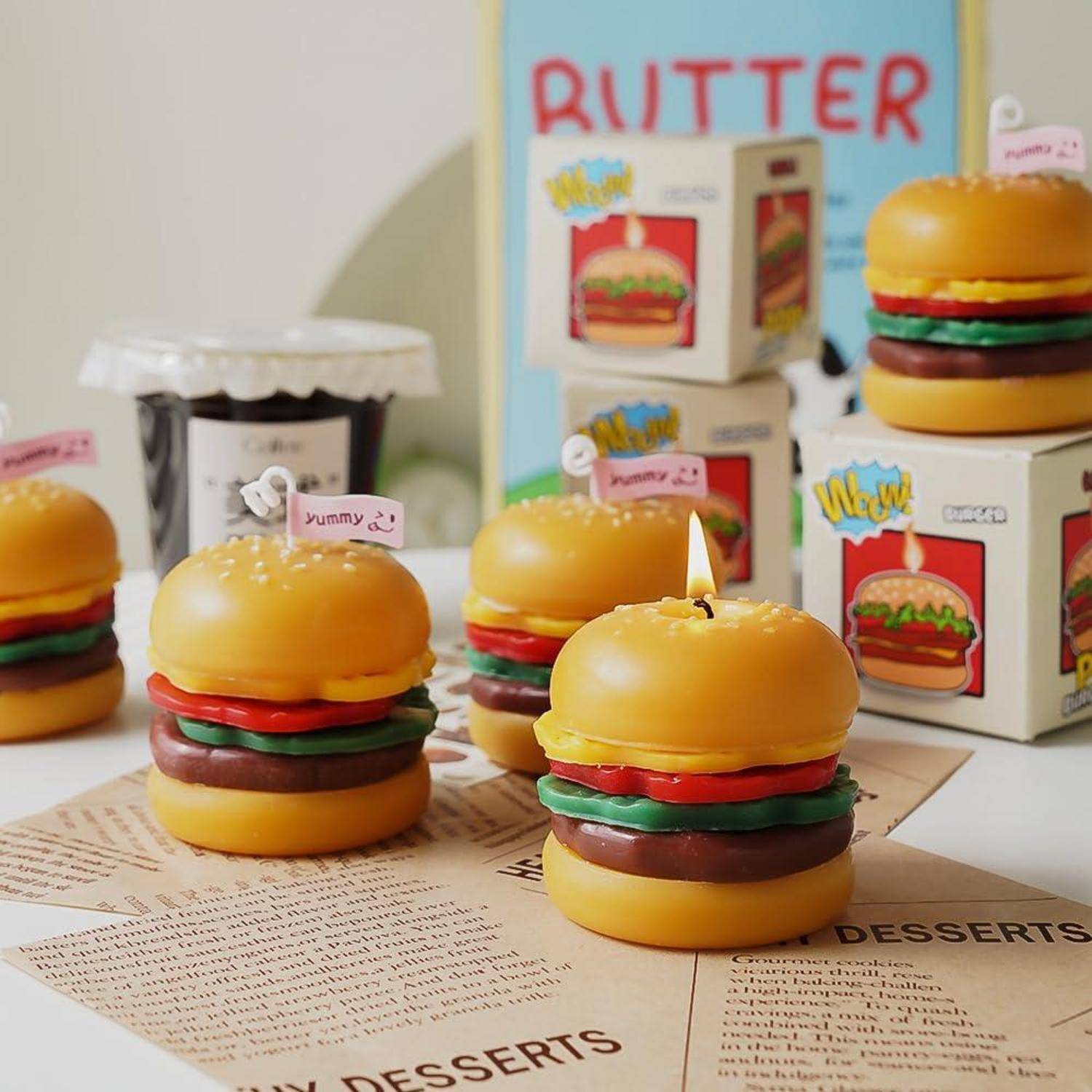 Miniatur-Burger-förmige Dessert-Kerzen auf einer Rezeptbuchseite, mit einem Butterschild und Dessert-Verpackung im Hintergrund.