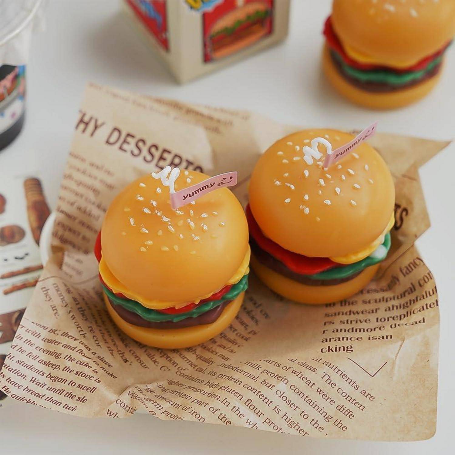 Kleine burgerfömige Nachtischsnacks mit sesamähnlichen Streuseln und Yummy-Tags, in Zeitungspapier eingewickelt auf einer weißen Oberfläche.