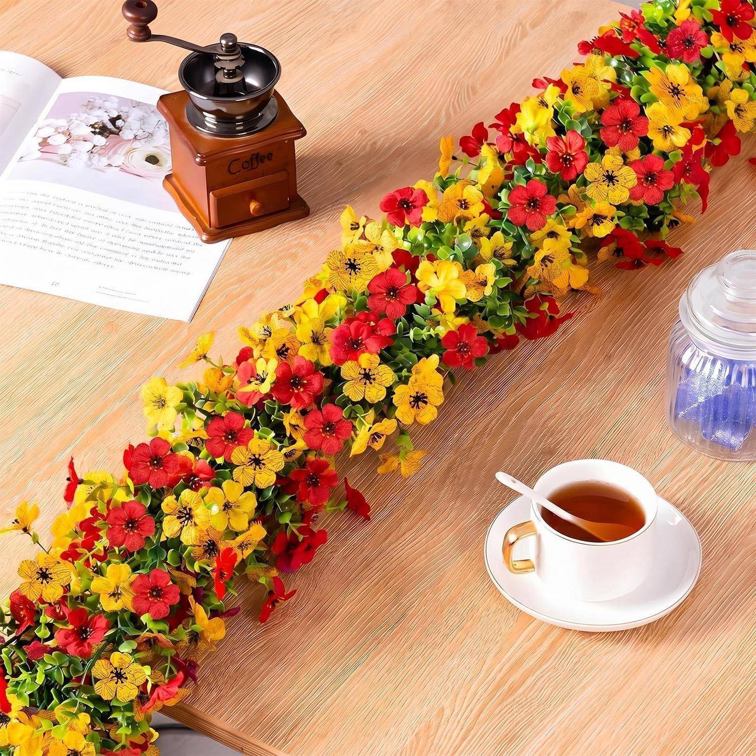 Lange Girlande aus roten und gelben Blumen über einem Holztisch verteilt, mit einer Kaffeetasse, einer Mühle, einem aufgeschlagenen Buch und einem Glasgefäß in der Nähe.