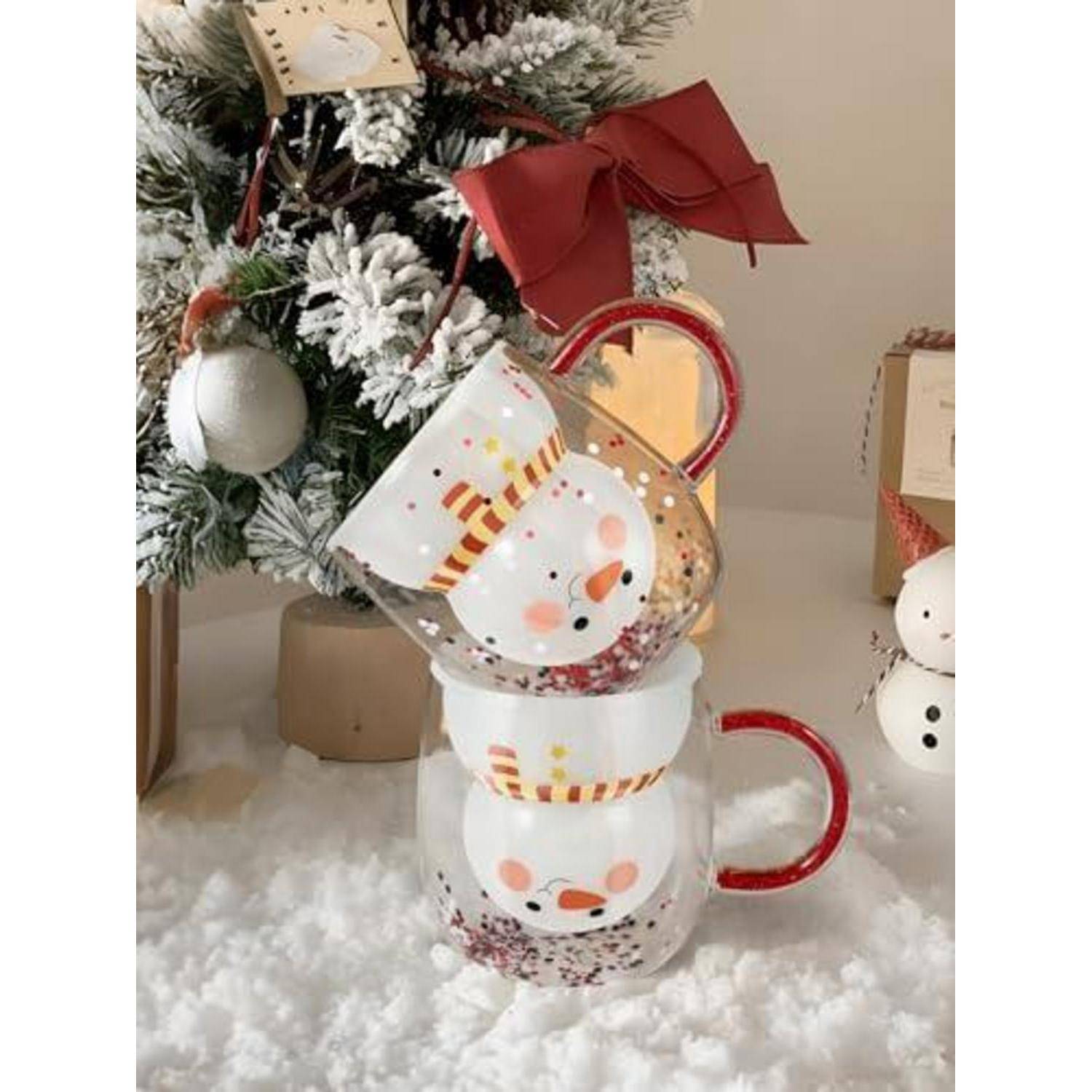 Gestapelte Schneemann-Tassen mit roten Henkeln neben einem geschmückten Weihnachtsbaum auf künstlichem Schnee.