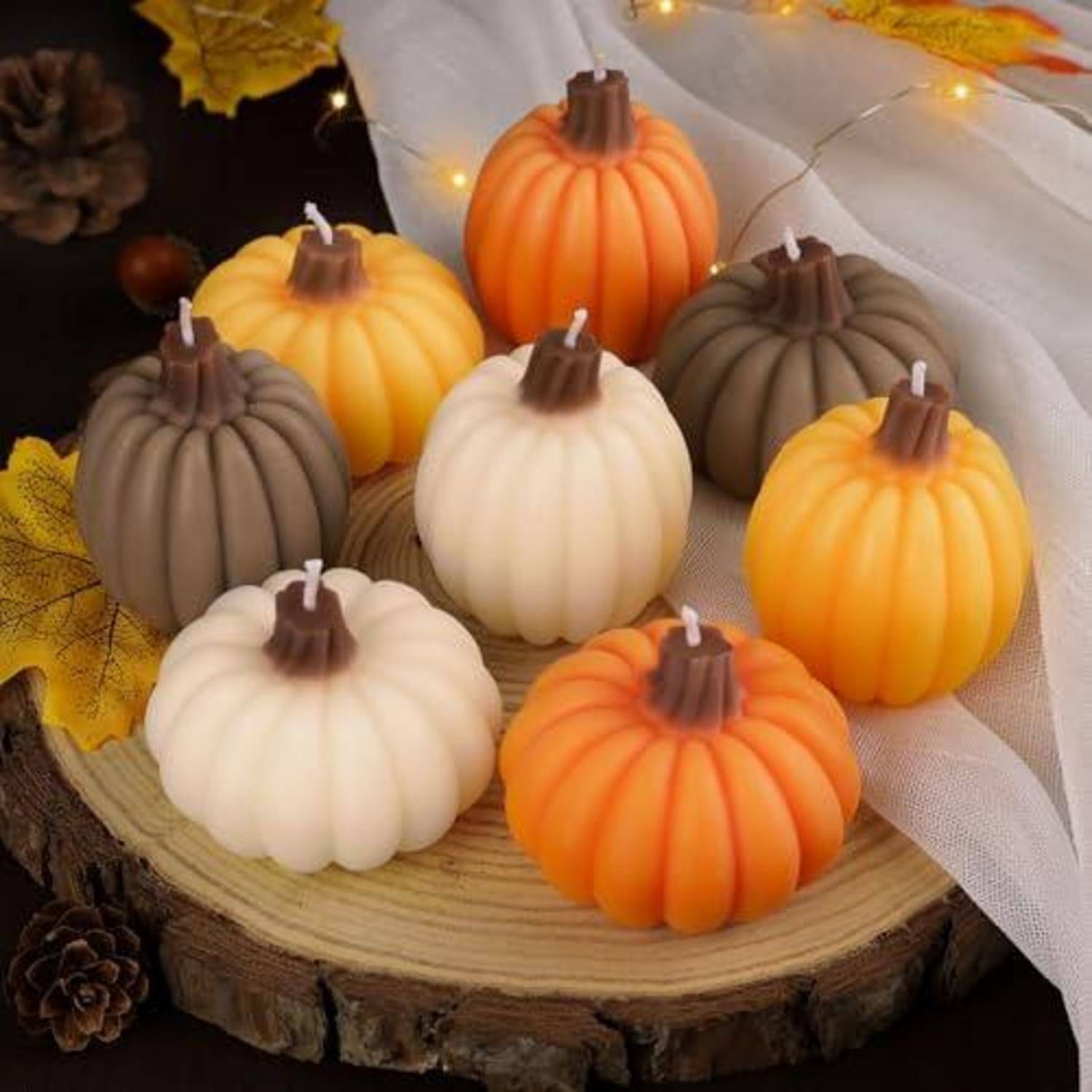 Verschiedene kürbisförmige Kerzen in Weiß, Orange, Gelb und Grau auf einer Holzplatte mit Herbstblättern und Lichterketten.