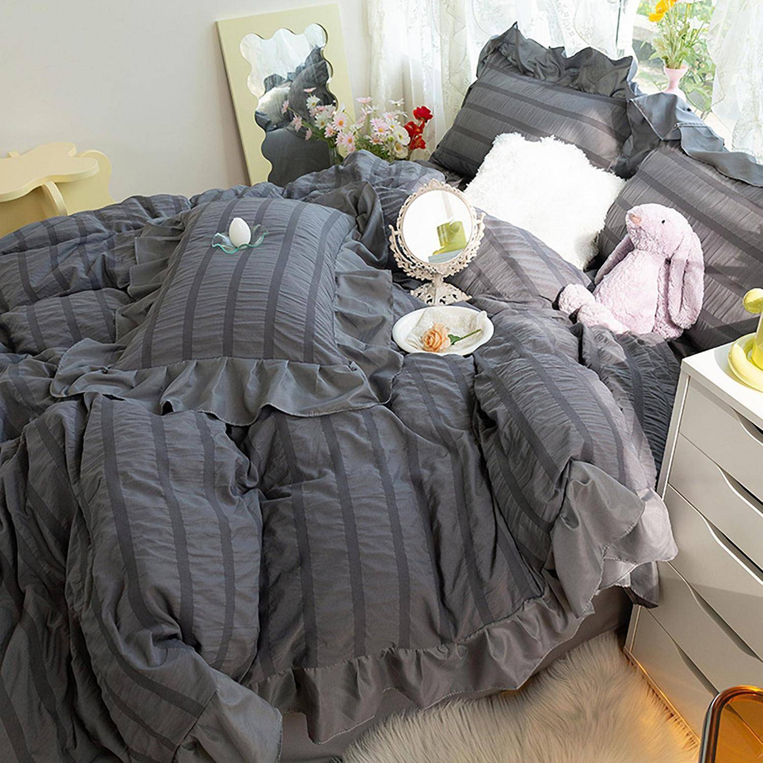 Ein ordentlich gemachtes Bett mit dunkelgrauem Rüschenbettzug, weißen Kissen, einem rosa Plüschhase und dekorativen Tabletts und Blumen in einem hellen Schlafzimmer.