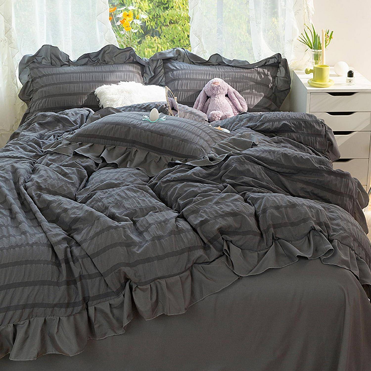 Ein ordentlich gemachtes Bett mit dunkelgrauer gerüschter Bettwäsche, zwei Kissen, einem weißen Wurf-Kissen und einem Lavendel-Häschen-Plüsch in einem hellen Schlafzimmer.