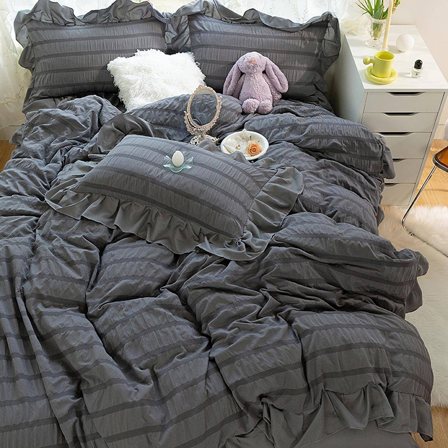 Ein ordentlich gemachtes Bett mit dunkelgrau gestreifter Bettwäsche, Kissen mit Rüschen, einem weißen flauschigen Kissen, einem lila Plüschtier und einem kleinen Nachttisch mit einer Pflanze und einer Tasse.