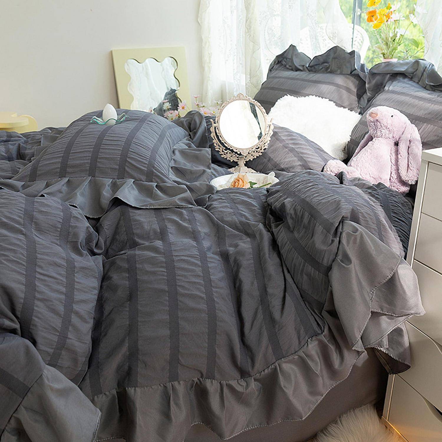 Ein ordentlich gemachtes Bett mit dunkelgrauem, gerafftem Bettzeug, weißen Kissen und dekorativen Spiegeln neben einem gefüllten lila Kaninchen in einem hellen Schlafzimmer.