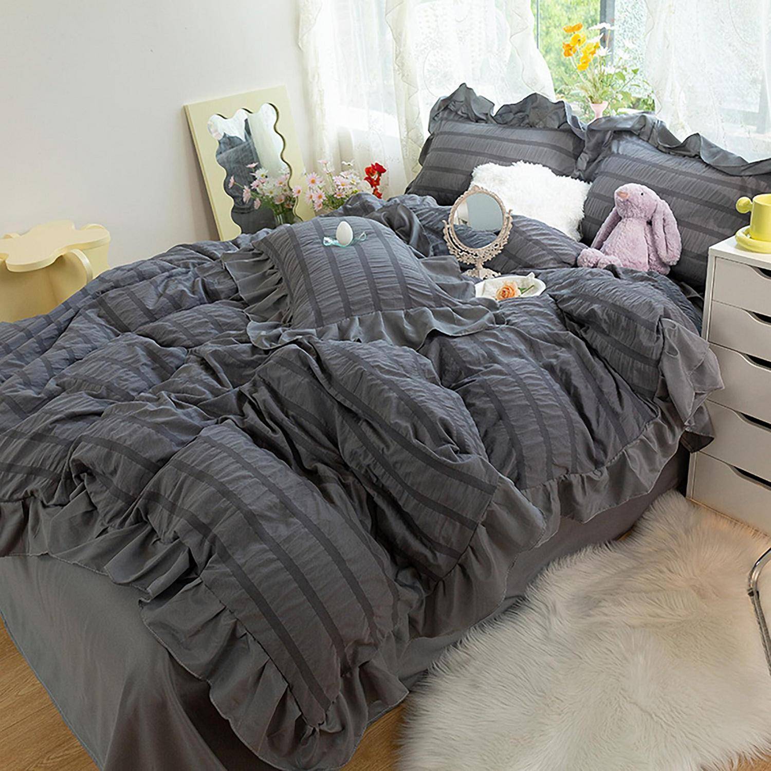 Ein ordentlich gemachtes Bett mit dunkelgrauem gerafftem Bettzug in einem hellen Schlafzimmer, mit Kissen, einem kleinen Spiegel, einem Plüschtier und einem Nachttisch.