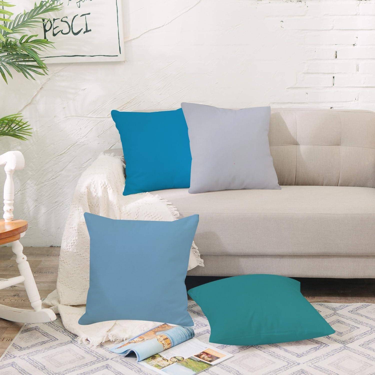Beiges Sofa mit vier blauen und grauen Dekokissen, einer weißen Decke und Zeitschriften auf einem gemusterten Teppich in einem hellen Wohnzimmer.