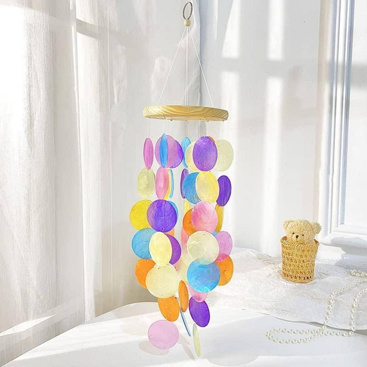Buntes hängendes Mobile mit durchsichtigen ovalen Scheiben, die von einem goldenen Ring in einem hellen, sonnendurchfluteten Zimmer herab baumeln, neben einem kleinen Teddybär in einem Körbchen.