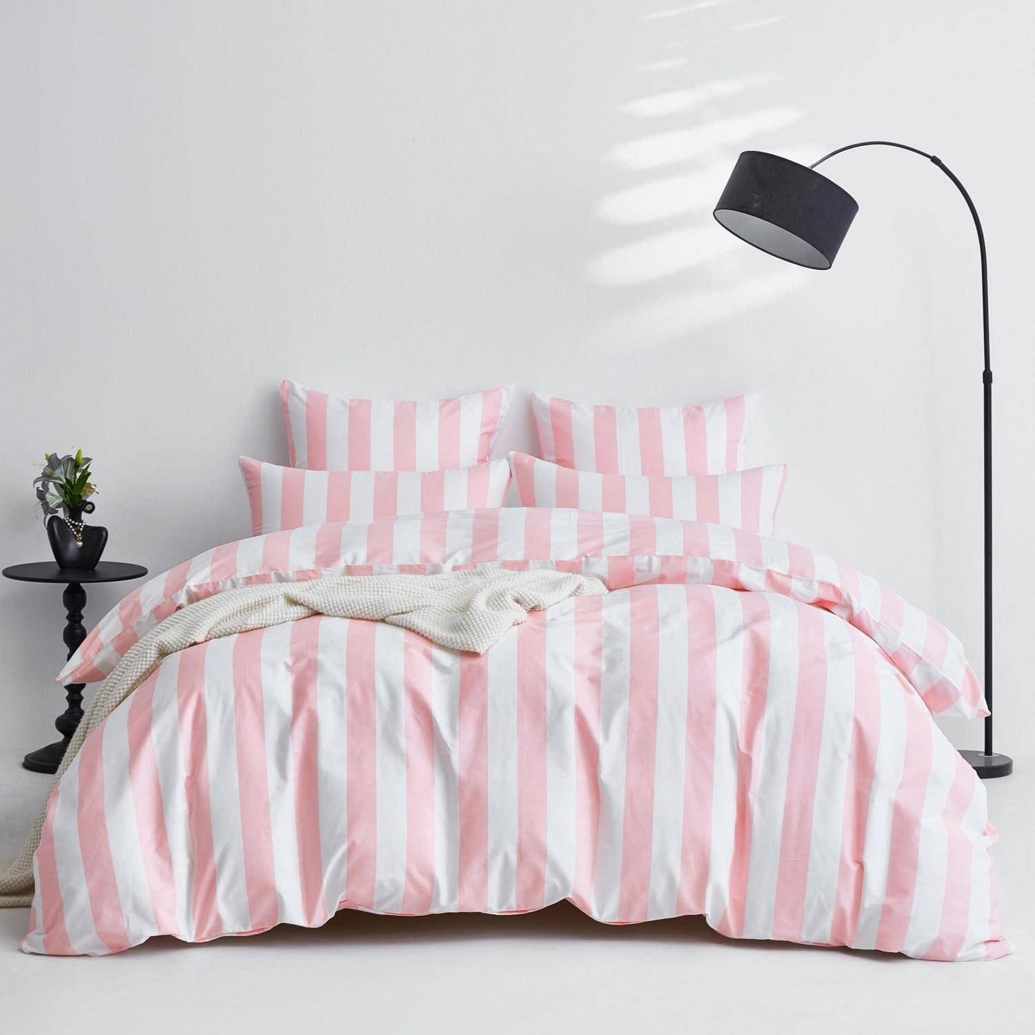 Rosa und weiß gestreiftes Bettzeugs auf einem ordentlich gemachten Bett, mit einer schwarzen Stehleuchte und einem kleinen Nachttisch in einem minimalistischen Schlafzimmer.
