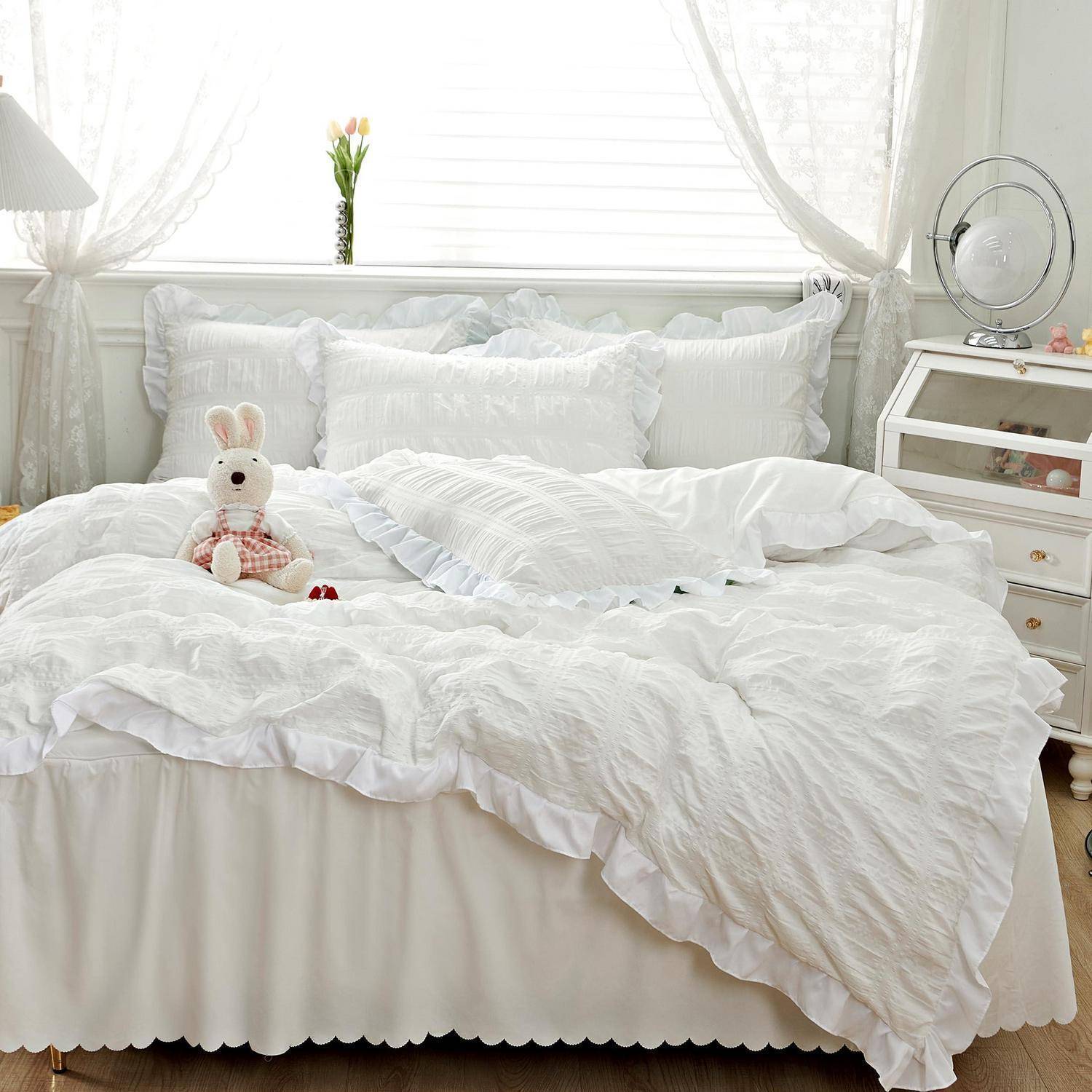 Schlafzimmer in hellem Weiß mit einem ordentlich gemachtem Bett, Kissen, einem Plüschhäschen, durchsichtigen Vorhängen und einem kleinen Ventilator und Tisch am Bettrand.