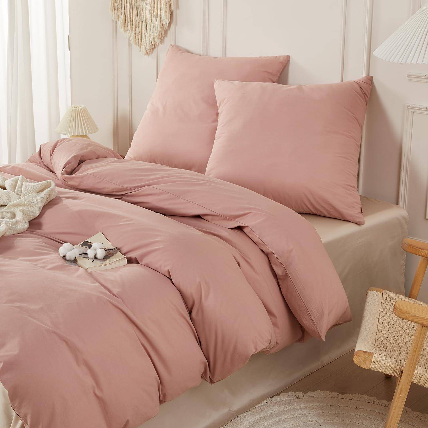 Ein Bett mit staubig rosa Bettwäsche und passenden Kissen in einem weichen, minimalistischen Schlafzimmer.