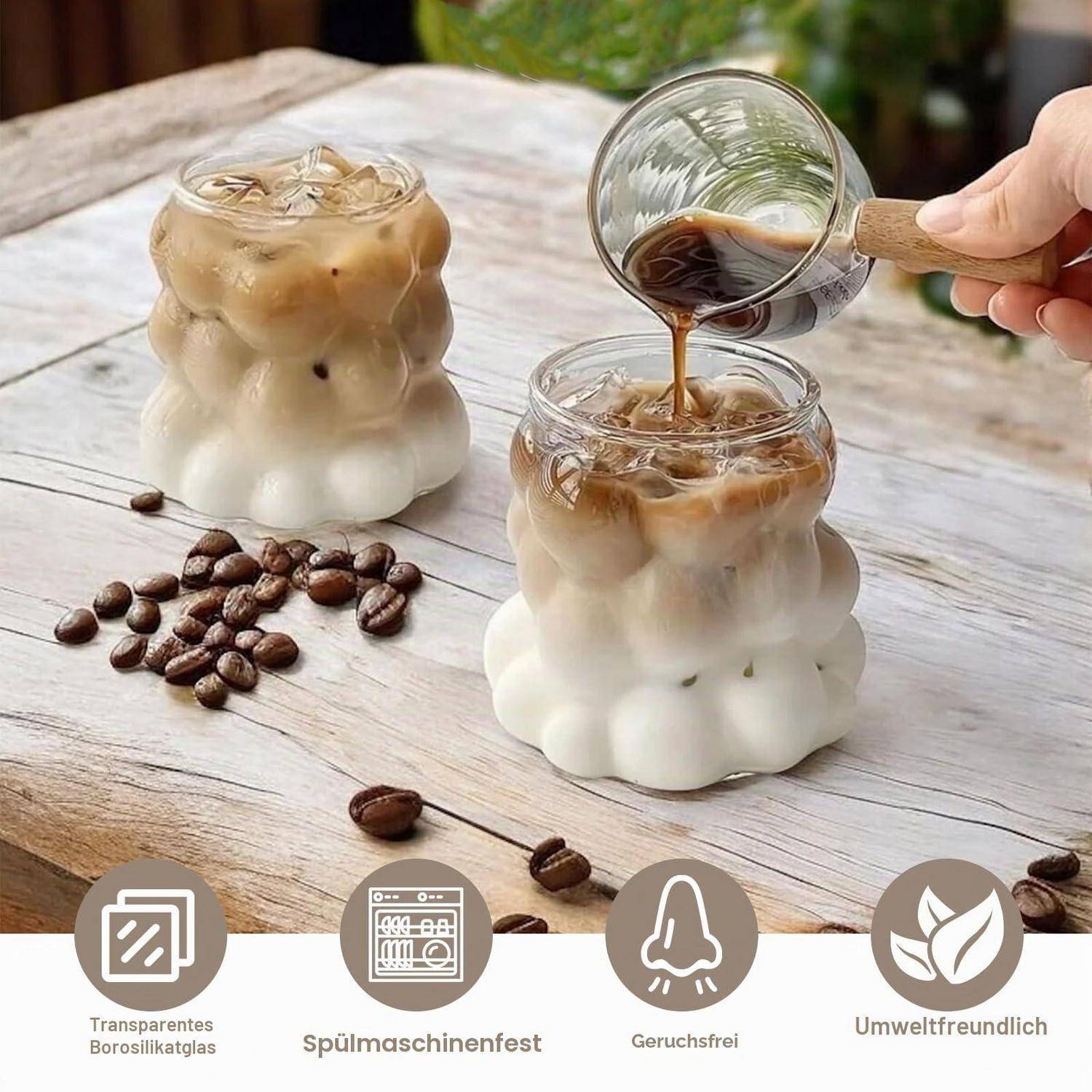 Zwei geschichtete Eiskaffee-Getränke in durchsichtigen Bubble-Gläsern, eines wird aus einer Glastasse eingegossen; Kaffeebohnen und deutsche Etiketten darunter werben für wiederverwendbare, geruchsneutrale, umweltfreundliche Tassen.