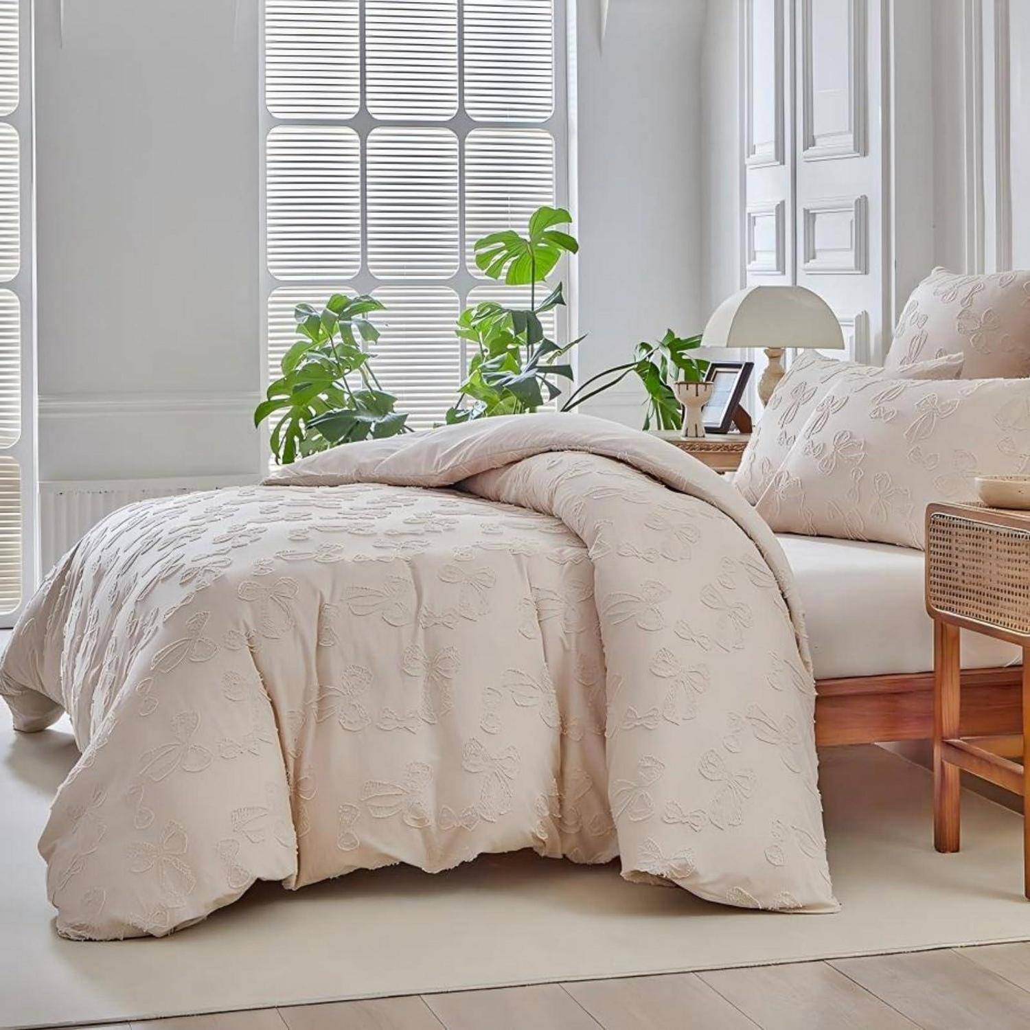Ordentlich gemachtes Bett mit beige strukturierter Bettwäsche in einem hellen Schlafzimmer, neben einem hölzernen Nachttisch, Fenster und Zimmerpflanzen.