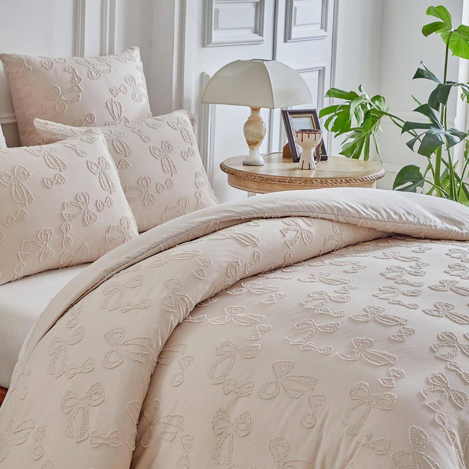 Beige Bettwäsche mit Blumenstickerei mit Kissen und einer zusammengefalteten Bettdecke in einem hellen Schlafzimmer neben einer Lampe, einem Beistelltisch und einer Pflanze.