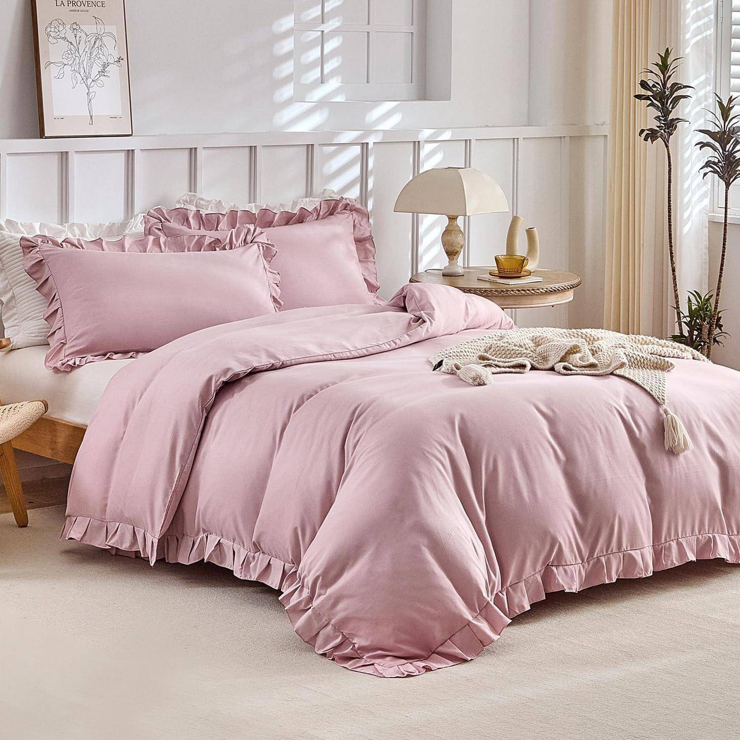 Rosa gerüschtes Bettwäsche-Set auf einem gemachten Bett in einem hellen, minimalistischen Schlafzimmer mit Nachttischlampe, Tablett und gerahmter Wandkunst.
