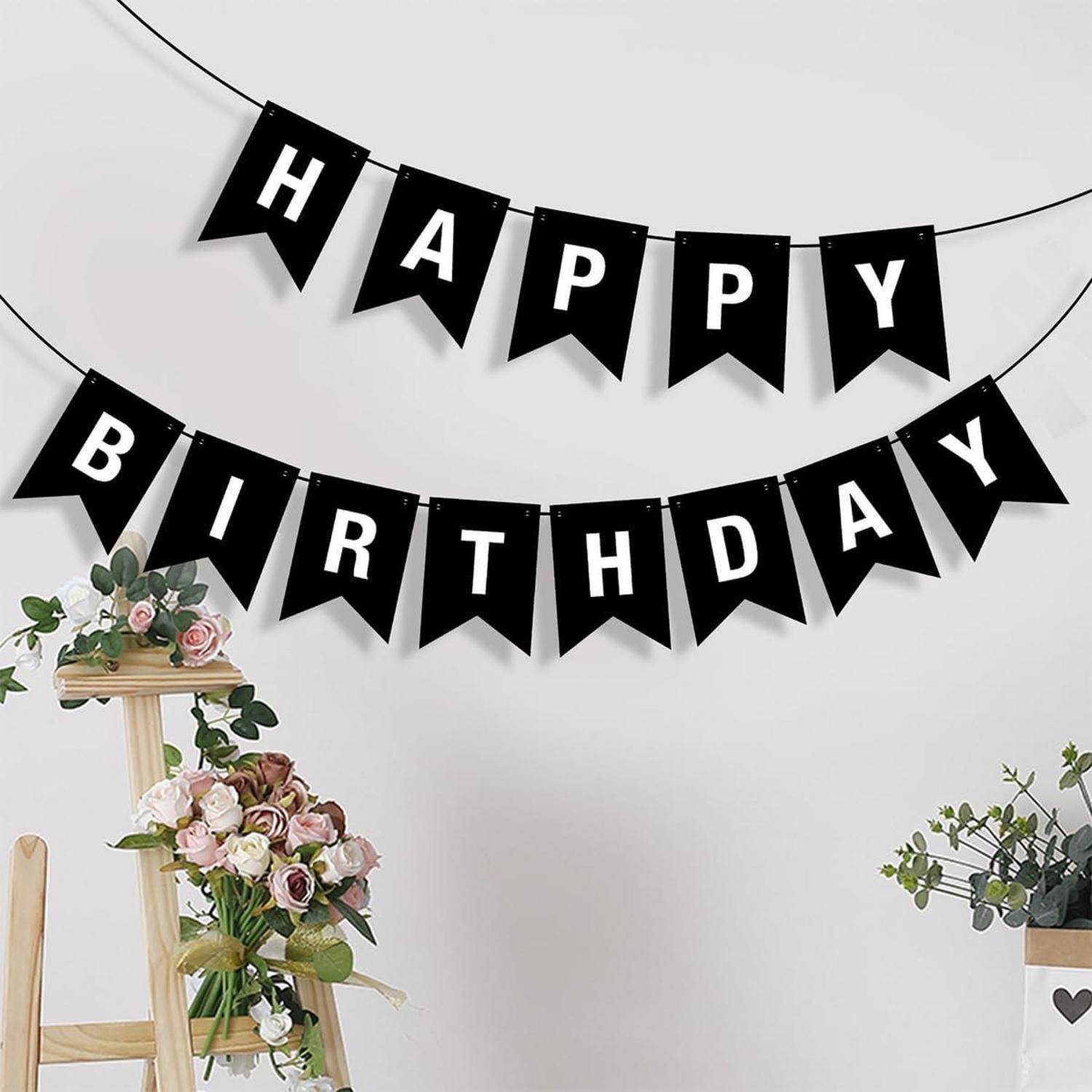 "Alles Gute zum Geburtstag"-Girlande in schwarzen Wimpelfahnen über floralen Dekorationen und einem Holzständer vor einer schlichten, hellen Wand.