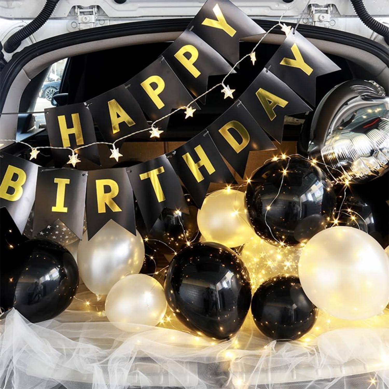 Schwarz-goldene Geburtstagsdekorationen mit "HAPPY BIRTHDAY""-Banner, Ballonen und Feenlichtern im Kofferraum eines Fahrzeugs.