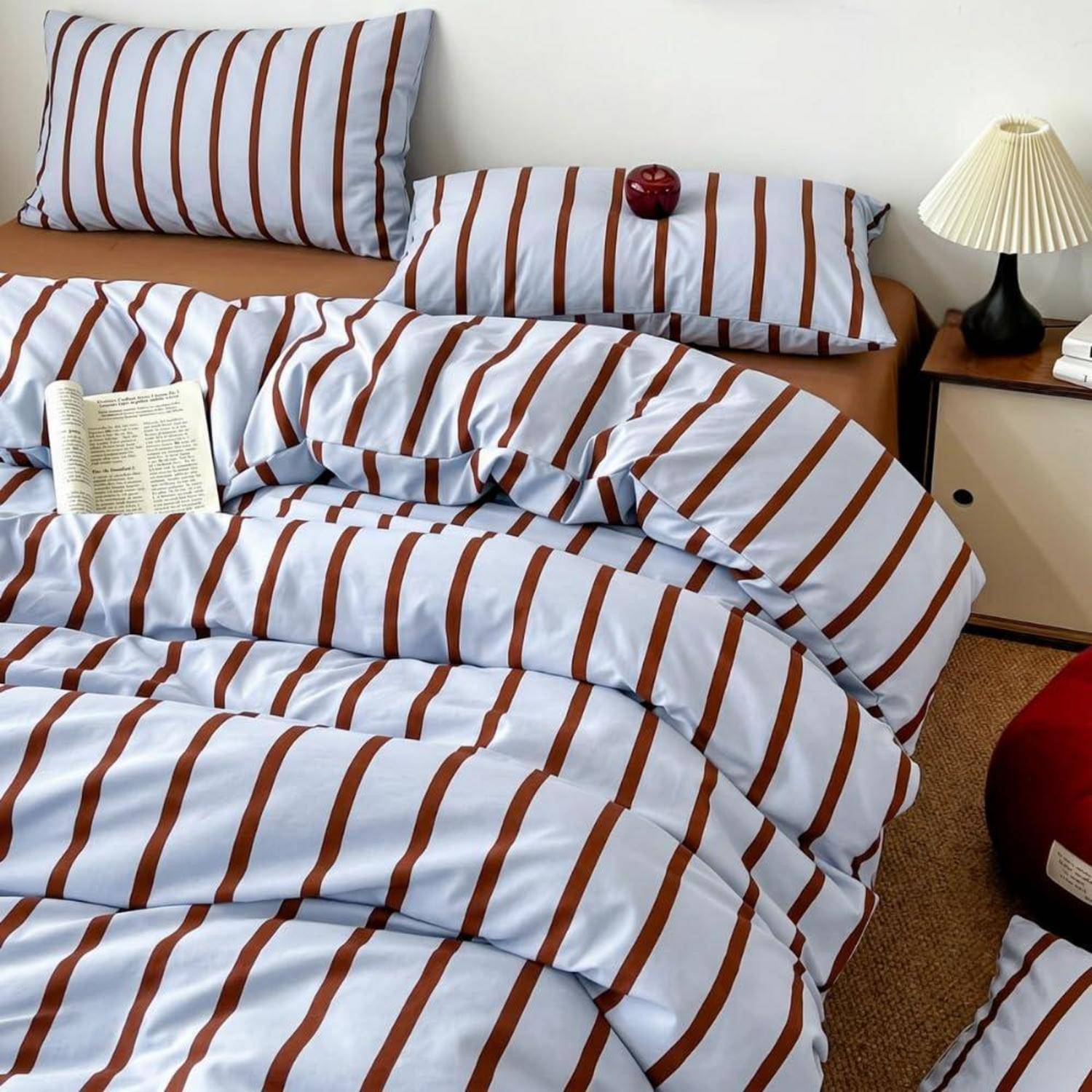 Ungemachtes Bett mit blau-braun gestreifter Bettwäsche, ein aufgeschlagenes Buch oben darauf, und ein Nachttisch mit einer Lampe und einem roten Apfel.
