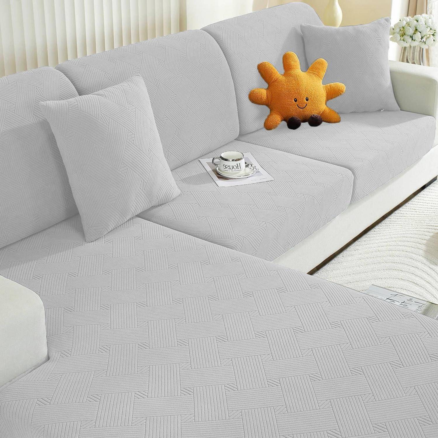Hellgrau kariertes Ecksofa mit strukturiertem Bezug, zwei passende Kissen, ein lächelndes orange Sonne-Plüschtier und eine Tasse auf einem Tablett in einem hellen Wohnzimmer.