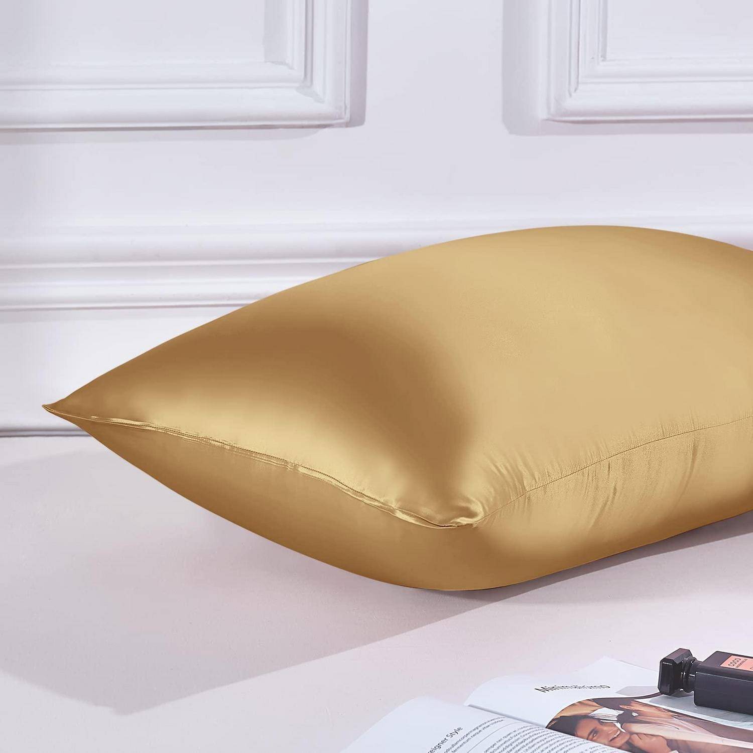 Goldsatin-Kissen auf einem weißen Bett, dargestellt in einem Close-up-Produktfoto mit einer teilweise sichtbaren Zeitschrift und einer kleinen Kamera im Vordergrund.