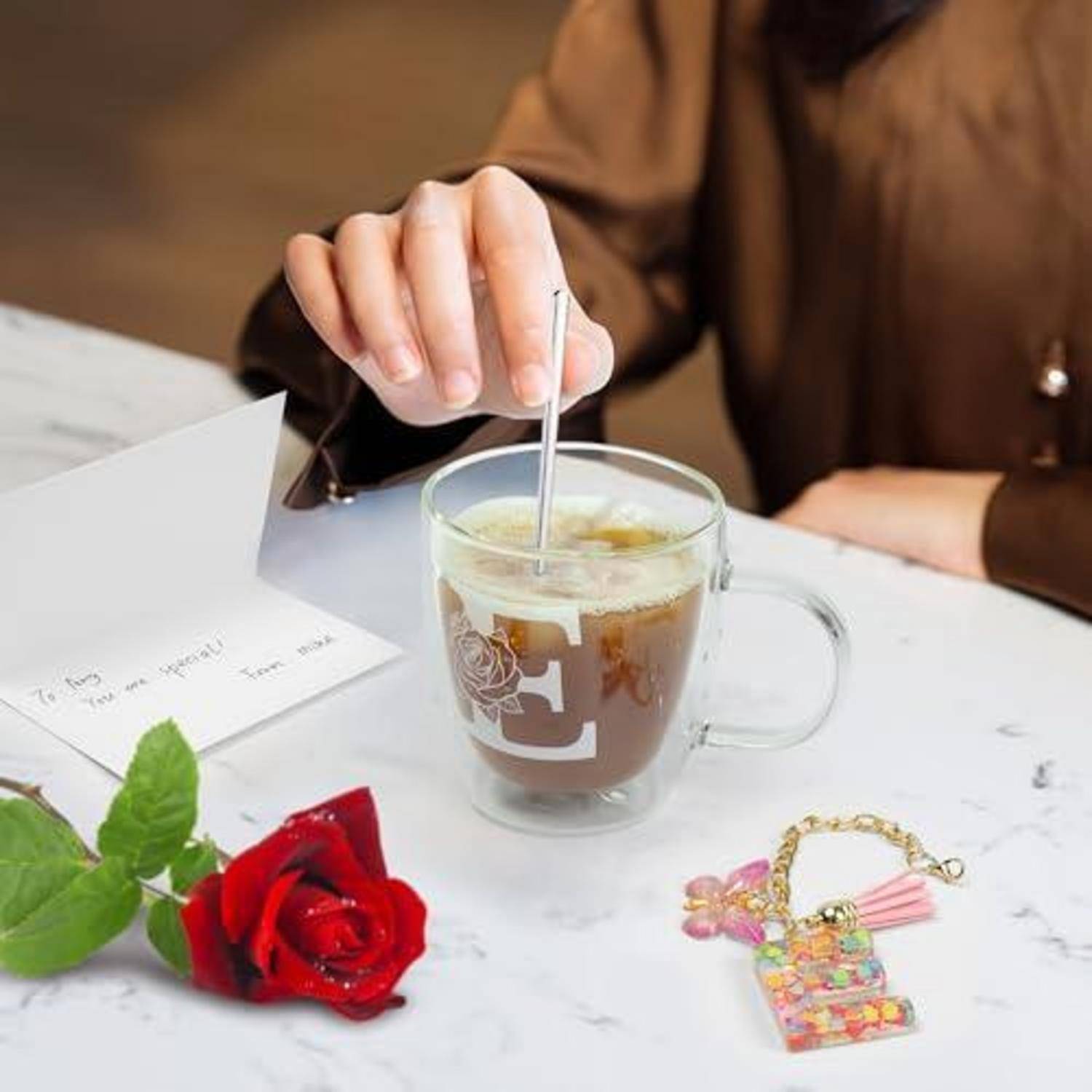 Person rührt ein Getränk in einem Glasbecher auf einem Tisch um, in der Nähe befinden sich eine rote Rose, eine Notizkartenund ein dekoratives Schlüsselband.