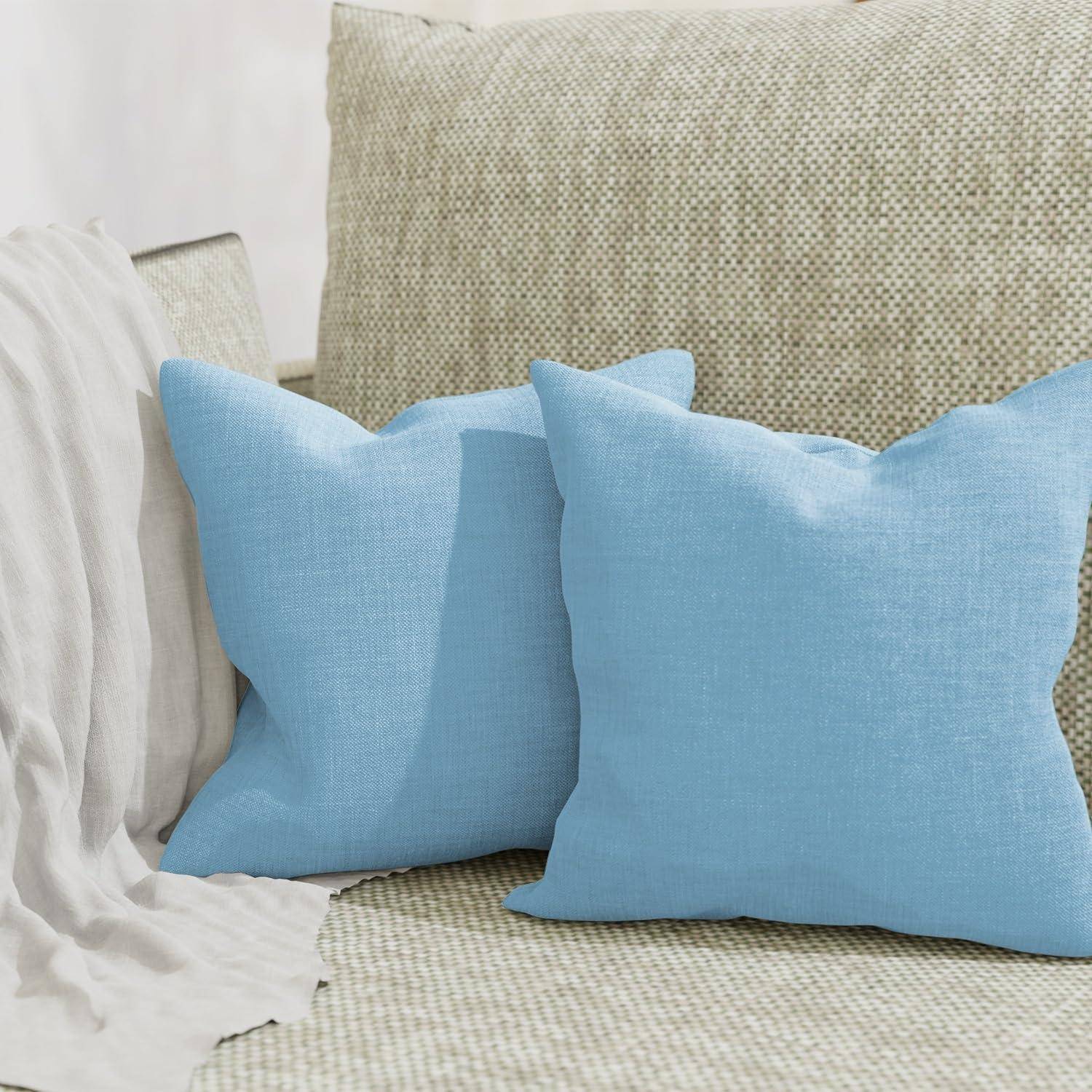 Zwei hellblaue Dekokissen auf einem beigen strukturierten Sofa neben einer drapierte weißen Decke.