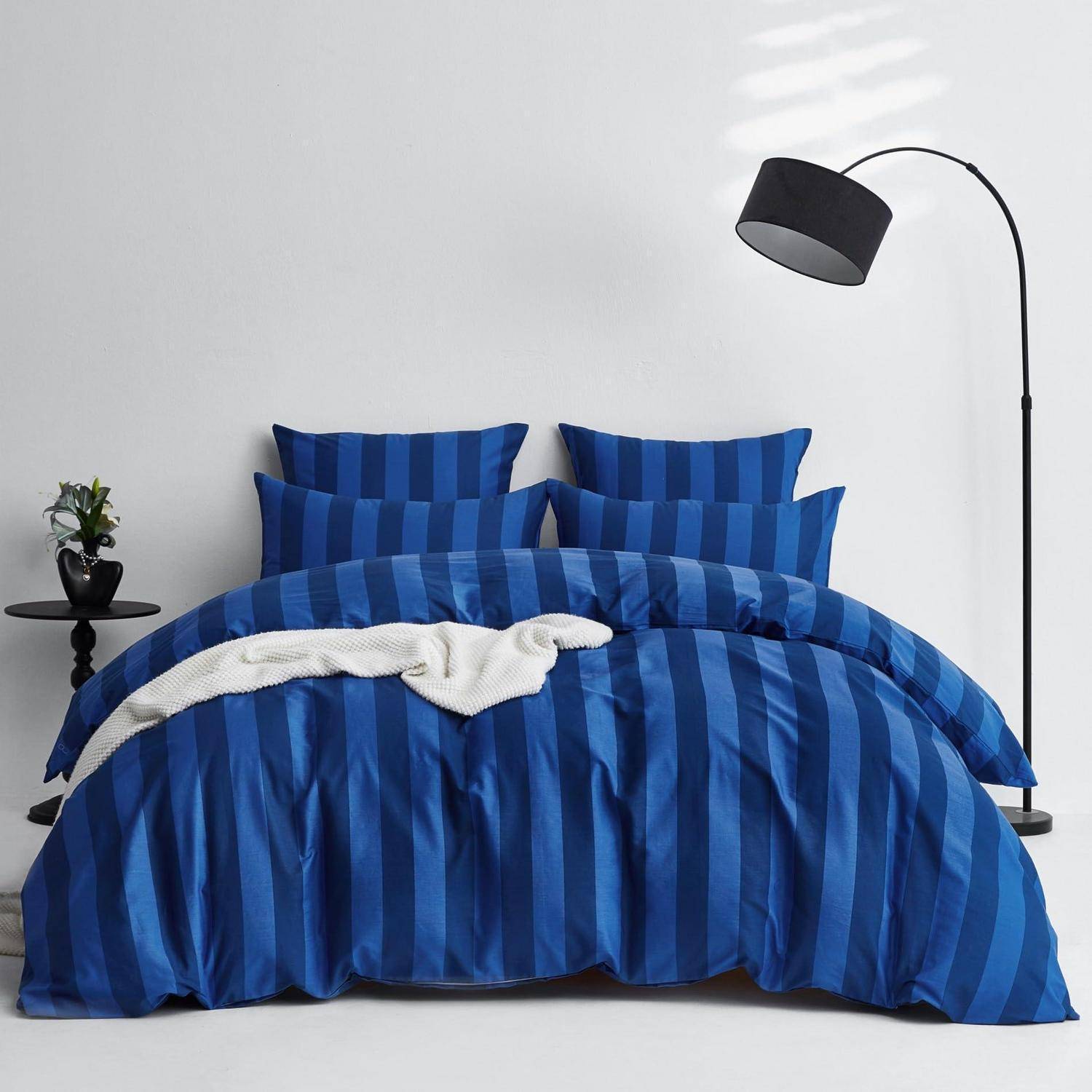 Eine blau gestreifte Tagesdecke und passende Kissen auf einem Bett, mit einer schwarzen Stehlampe und einem kleinen Beistelltisch an einer schlichten Wand.