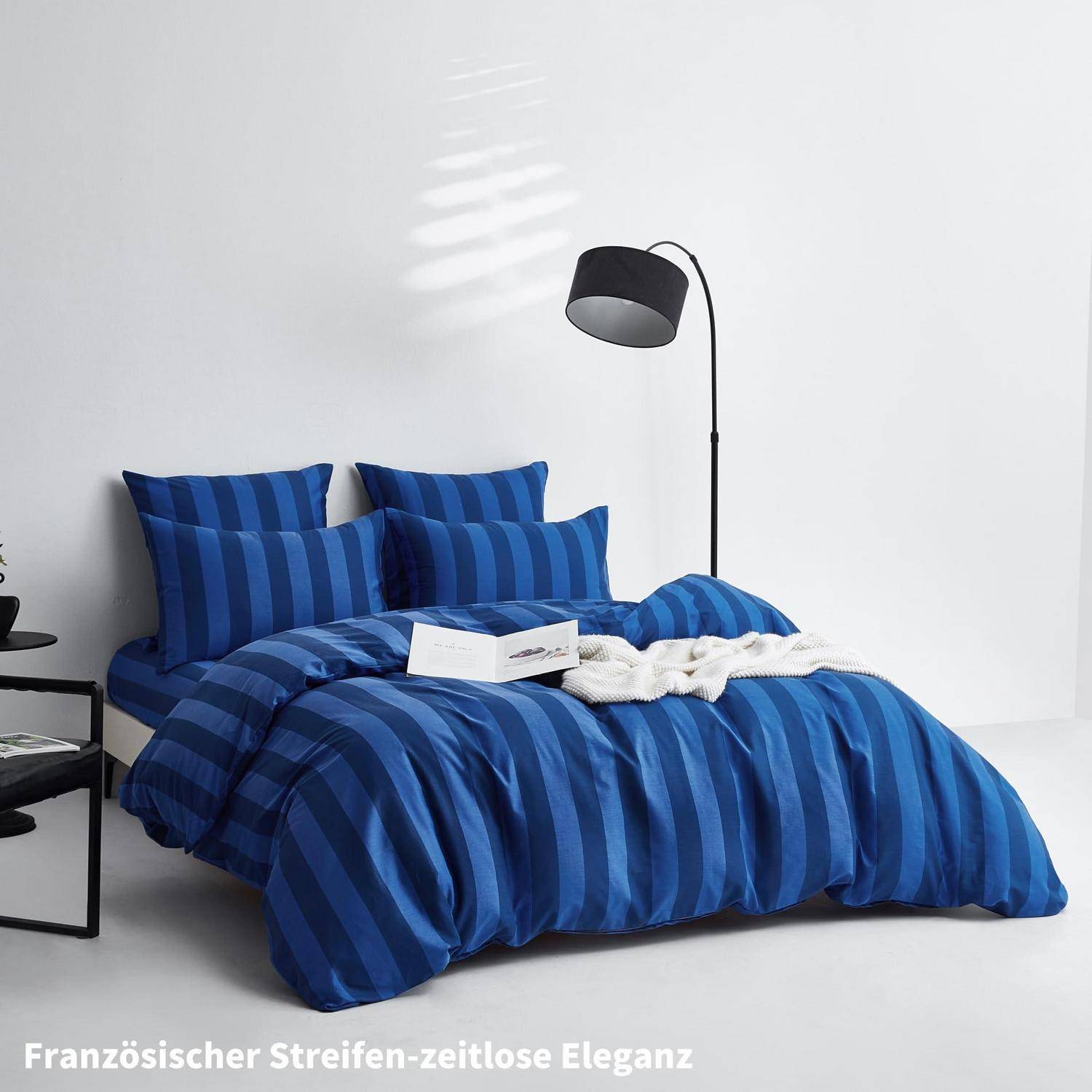 Bettwäscheset mit blauen Streifen auf einem Bett mit Kissen, Decke und einer Stehleuchte in einem minimalistischen weißen Schlafzimmer; der Text lautet „Französischer Streifen – zeitlose Eleganz".