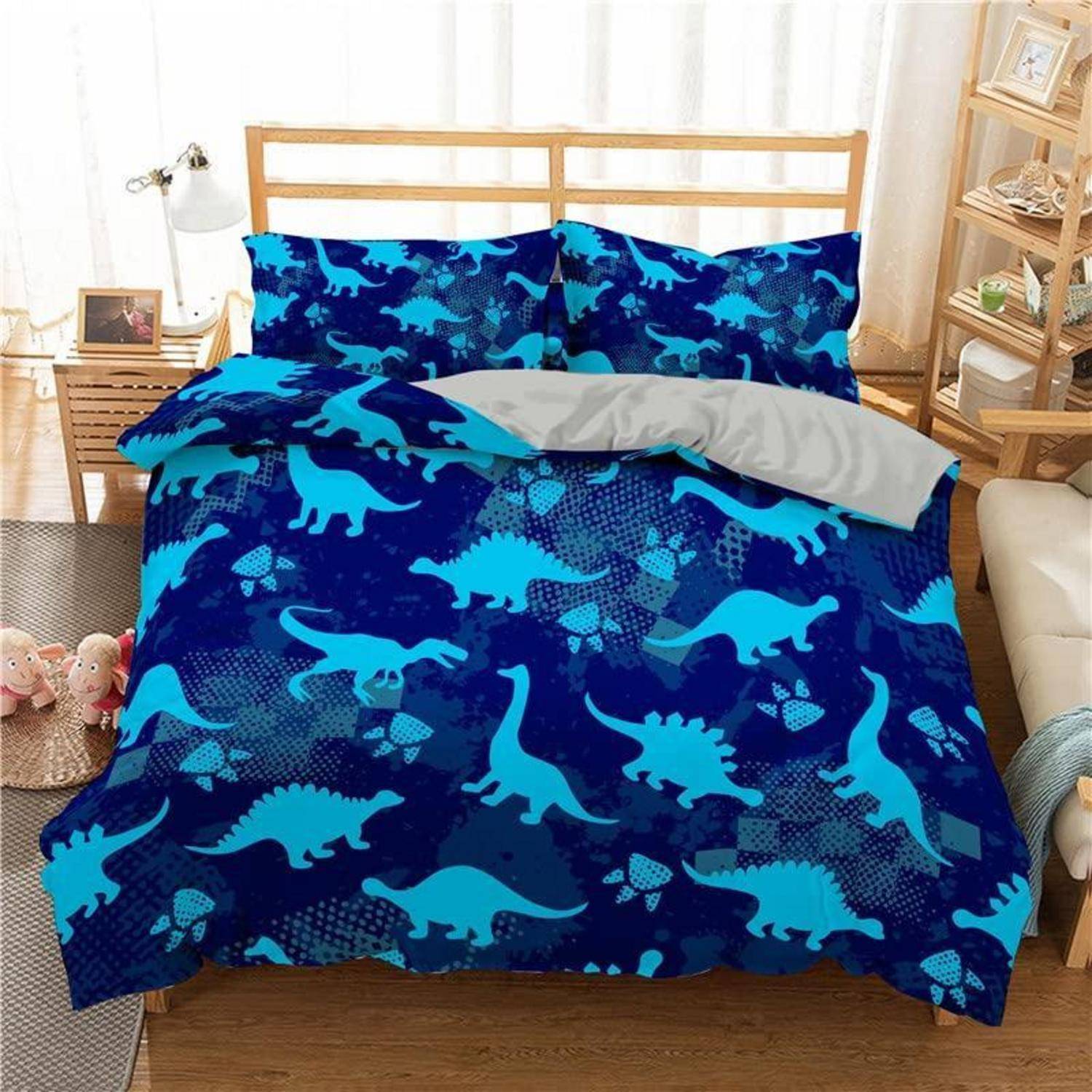 Bett mit blauem dinosauriermuster-Bettwäsche und passenden Kissenbezügen in einem hellen Schlafzimmer, mit einer grauen Wurfdecke oben übergefaltet.