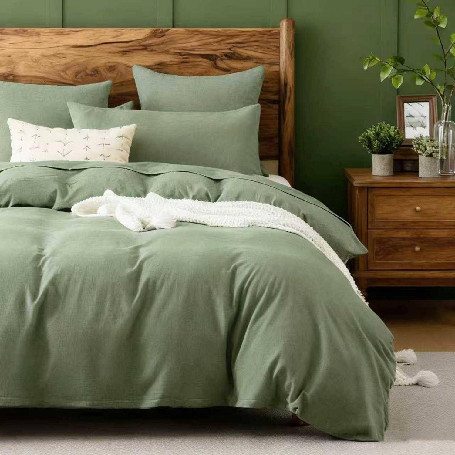 Ein ordentlich gemachtes Bett mit graugrüner Bettwäsche, weißer Wurfdecke und hölzernem Kopfteil neben einem Nachttisch mit Pflanzen in einem Schlafzimmer mit grünen Wänden.
