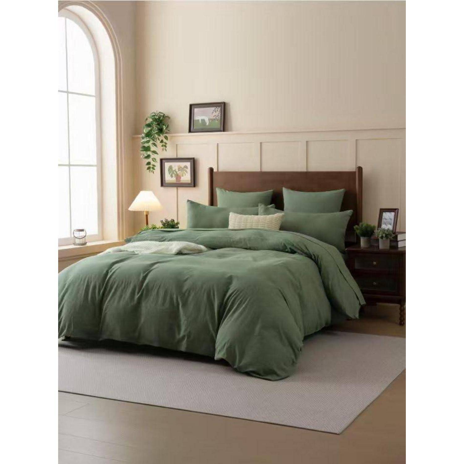 Ein ordentlich gemachtes Bett mit olivgrüner Bettwäsche steht in einem hellen Schlafzimmer neben einem großen gewölbten Fenster, einer Lampe, Nachttischen und Wandkunst.