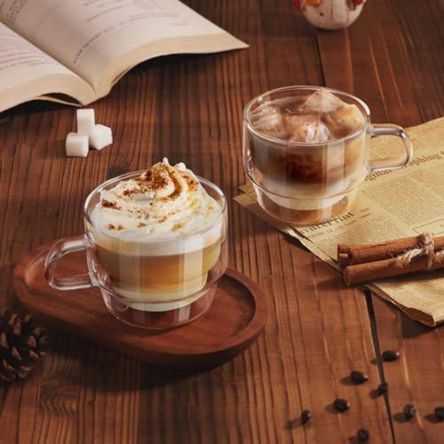 Zwei Glastassen mit geschichteten Kaffegetränken, die mit Schlagsahne gekrönt sind, stehen auf einem Holztisch, in der Nähe von Zimtstangen, Zuckerwürfeln und einem offenen Buch.
