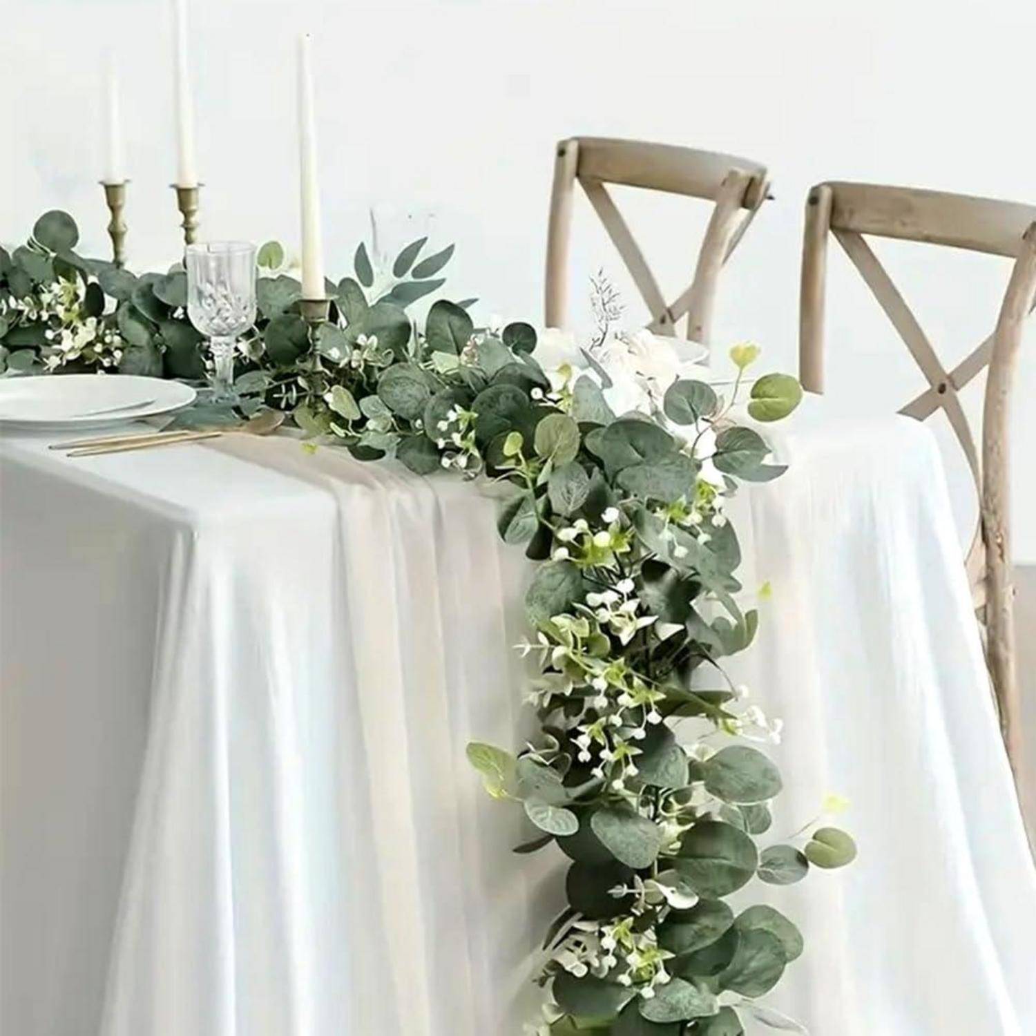 Eleganter Esstisch mit weißen Tischdecken, Kerzen, Glaswaren und einer über die Kante drapierten Tischmitte aus Eukalyptus und weißen Blüten.