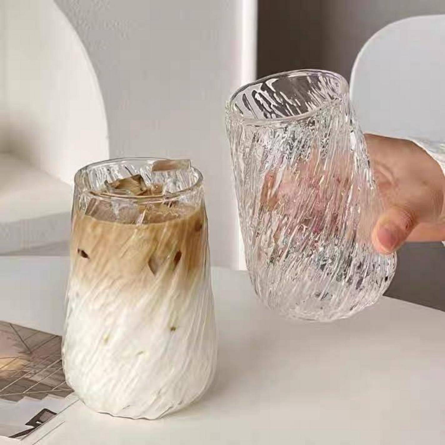 Zwei strukturierte klare Gläser nebeneinander gezeigt, eines gefüllt mit Eiskaffee, das andere leer in einer Hand gehalten.
