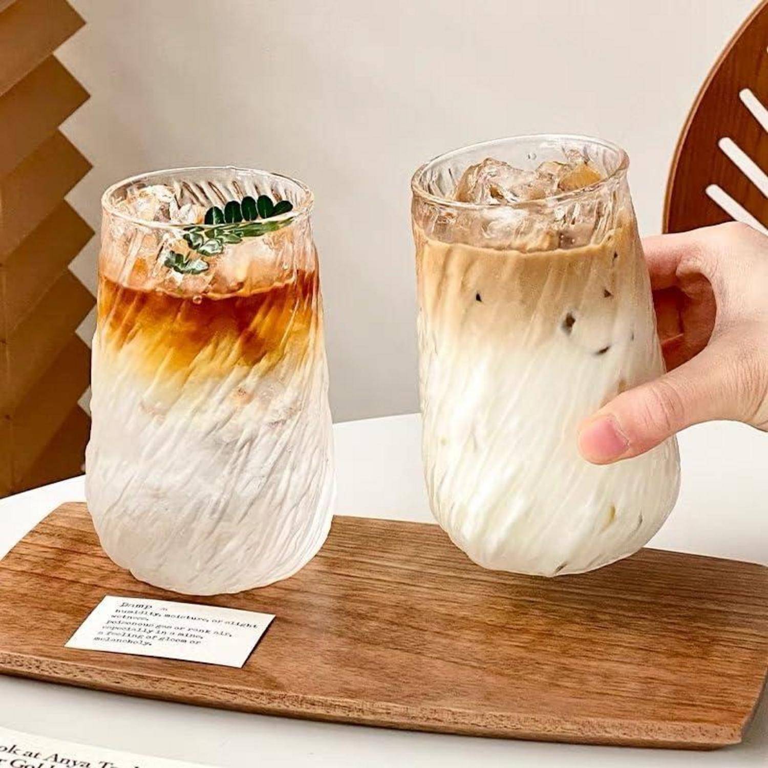 Zwei gefrorene geschichtete Kaffeegetränke in klaren Gläsern auf einem Holztablett, eines wird von einer Hand gehalten.