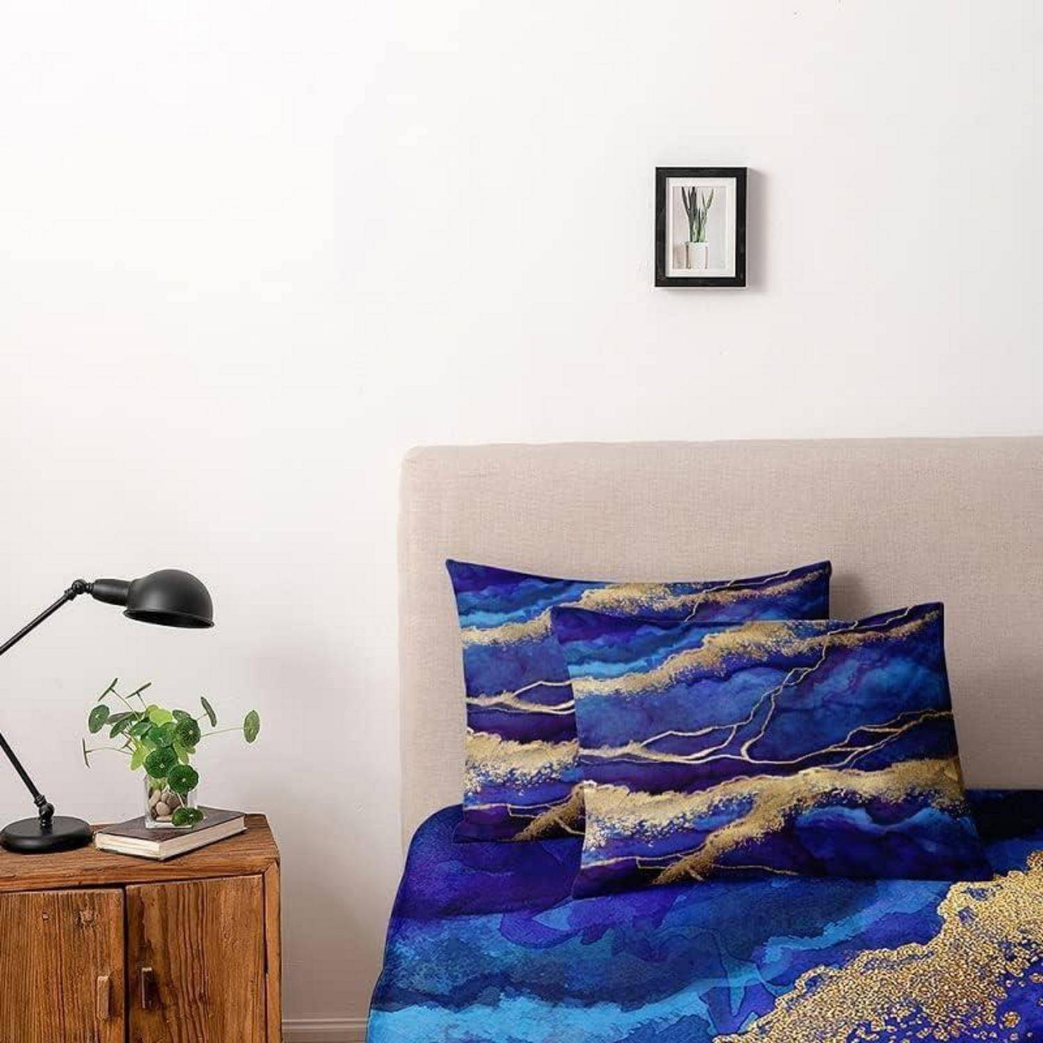 Ordentlich gemachtes Bett mit beigem Kopfteil und blau-lila gemusterter Bettwäsche, neben einem Holznachttisch mit Lampe und Pflanze, unter einem kleinen gerahmten Druck.