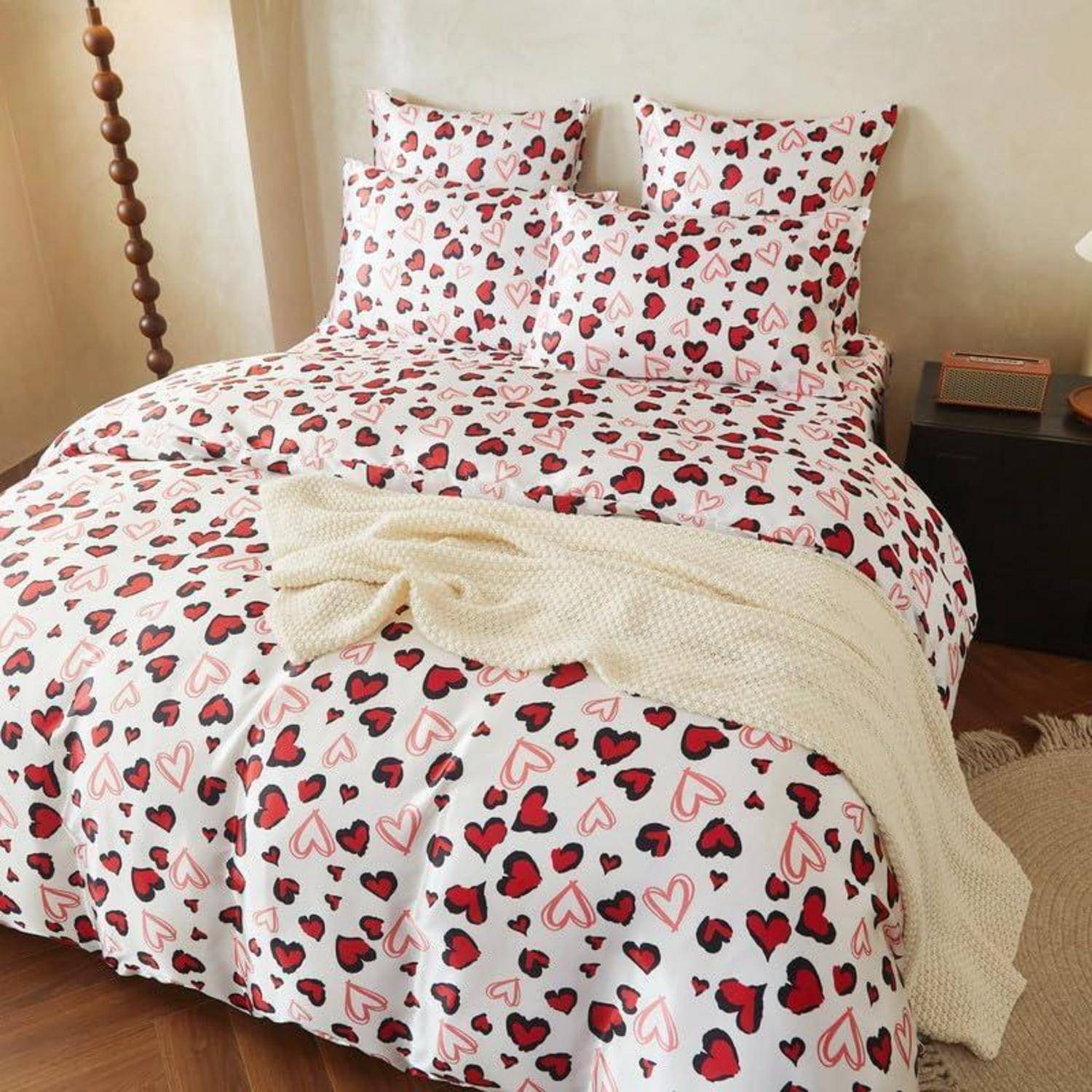 Bett mit weißer Bettwäsche und Kissen mit Herzmustern, einer cremefarbenen Wurfdecke, in einem warmen Schlafzimmer mit Nachttisch und Teppich.