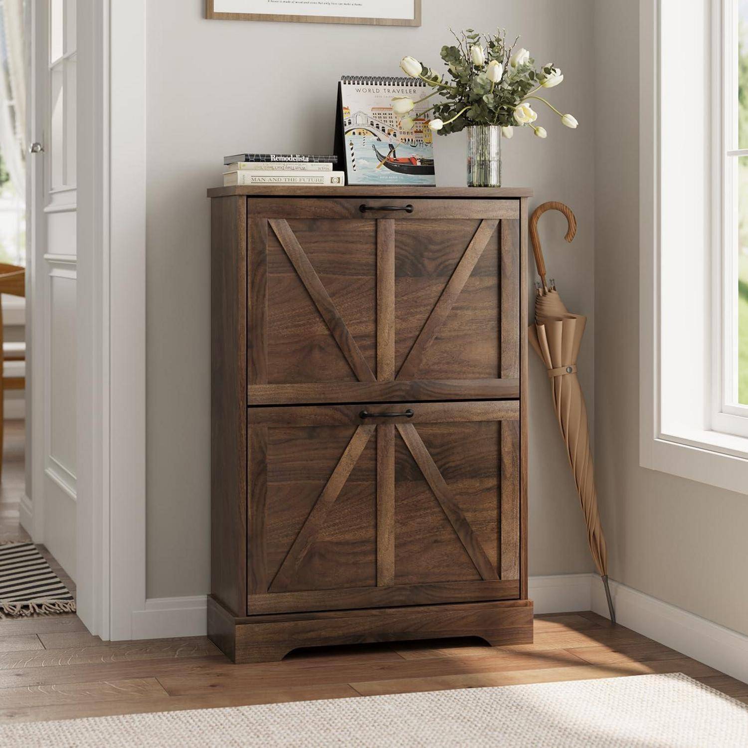 Holzschrank mit X-Muster-Türen, in dem Bücher, ein Kunstdruck im Rahmen, eine Blumenvase und ein Regenschirm neben einem hellen Fenster stehen.