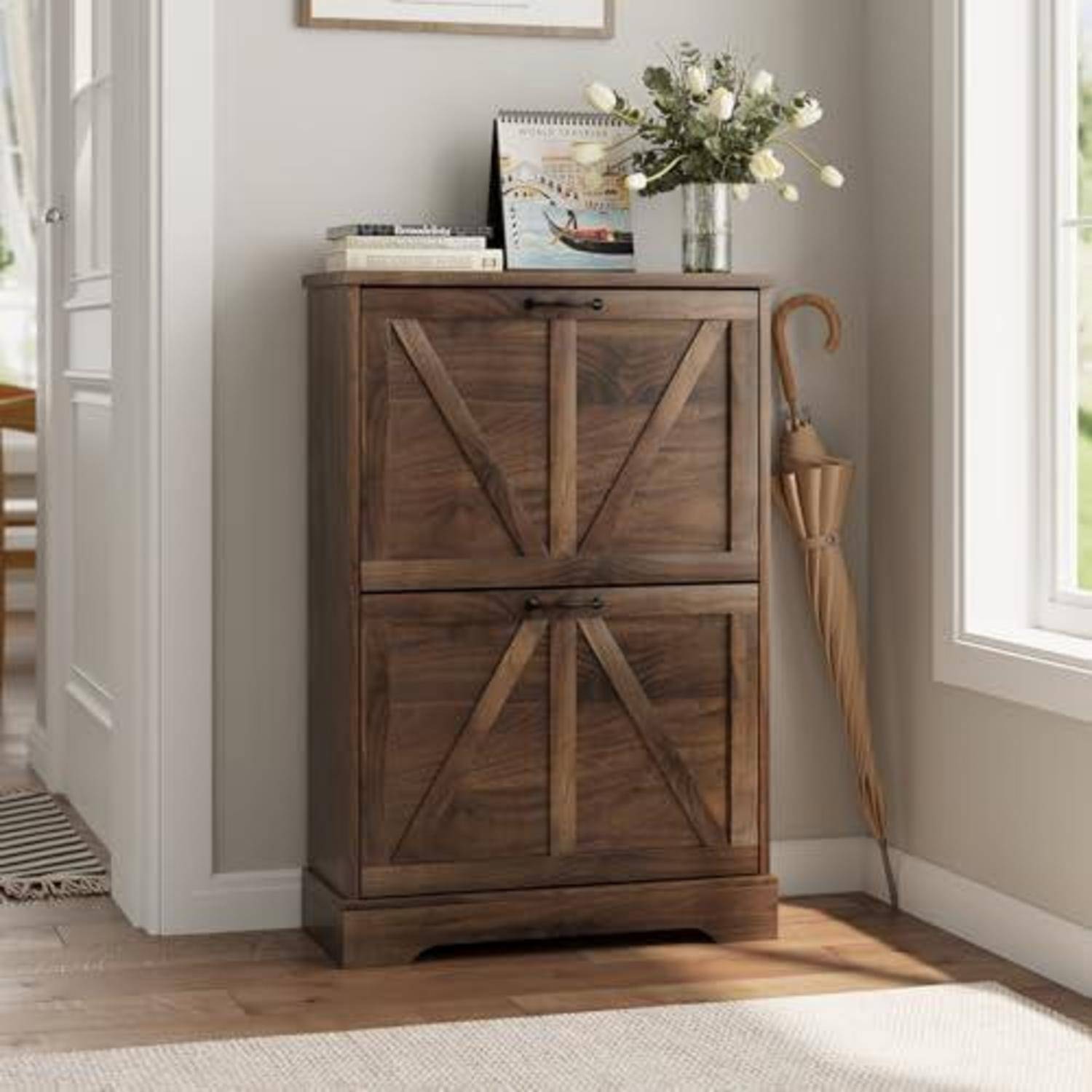 Holzschrank mit zwei Türen steht neben einem Fenster, oben beladen mit Büchern, gerahmten Kunstwerken und einer Vase mit Blumen, daneben lehnt ein Regenschirm.
