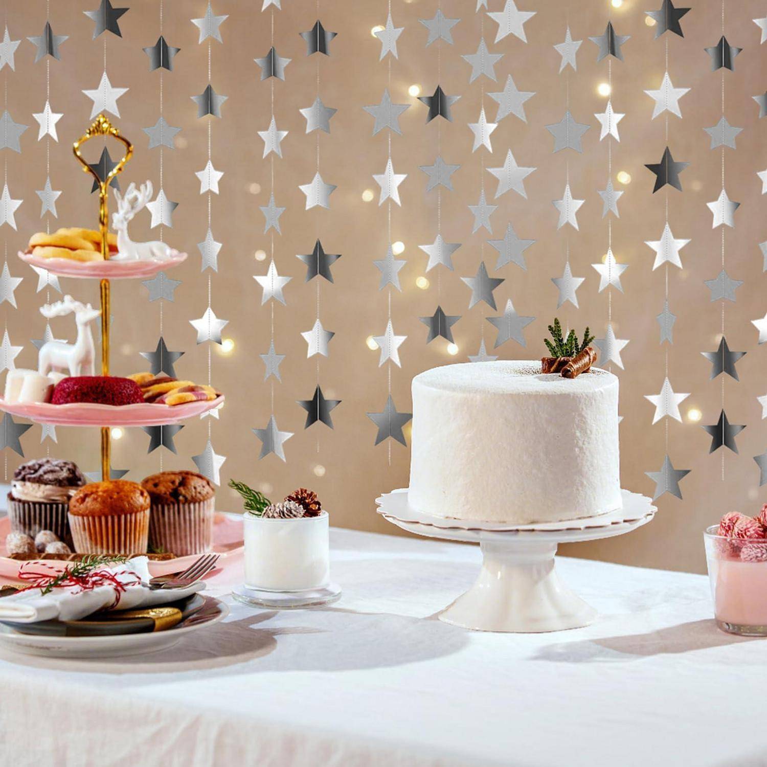 Weiße gefrostete Torte auf einem Sockel neben einem Dessertständer mit Gebäck und Cupcakes, auf einem Tisch vor einem silbernen Sternenhintergrund aufgestellt.