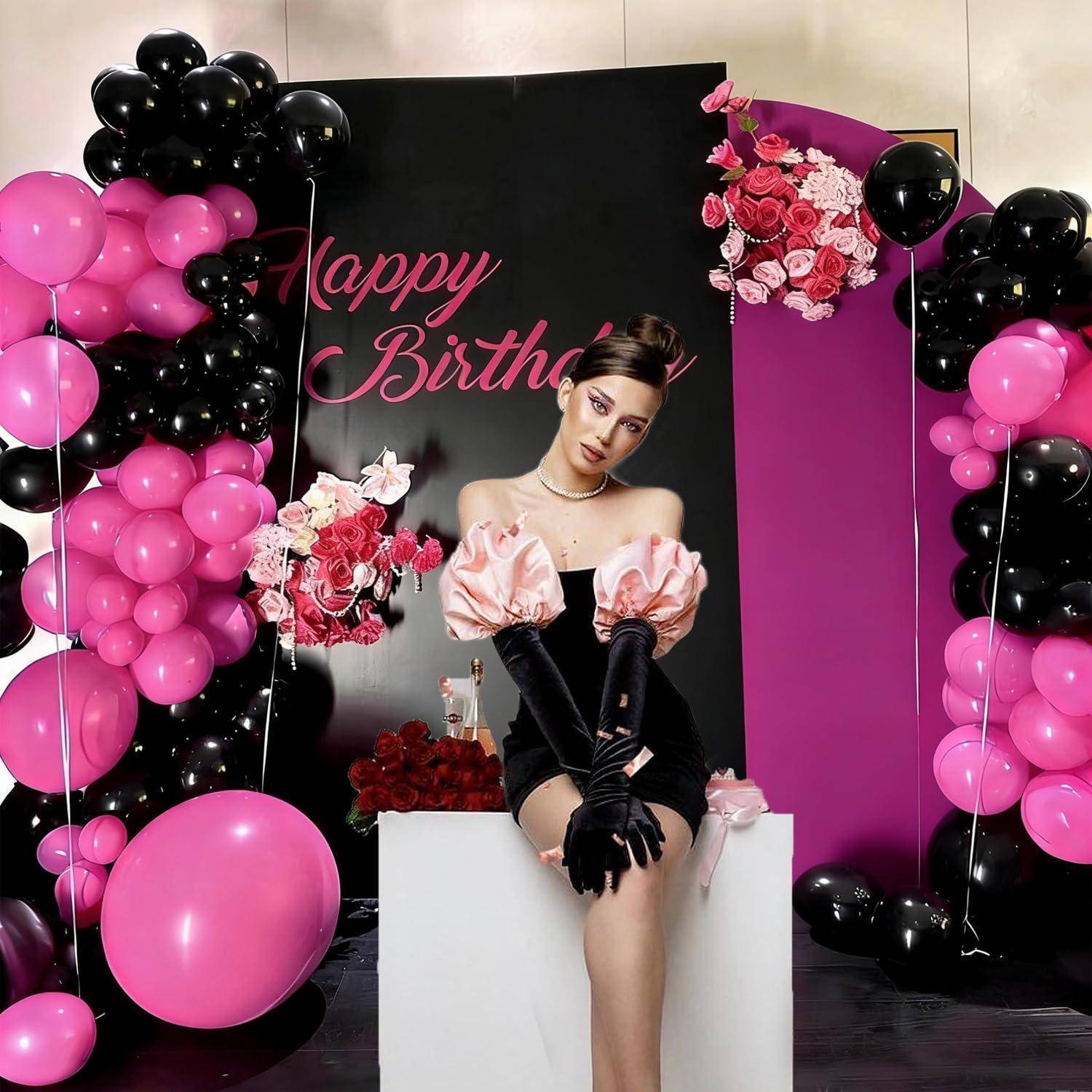 Eine Frau sitzt vor einer Geburtstagskulisse mit dem Text „Happy Birthday" und ist umgeben von rosa und schwarzen Luftballons sowie rosa Blumenarrangements.