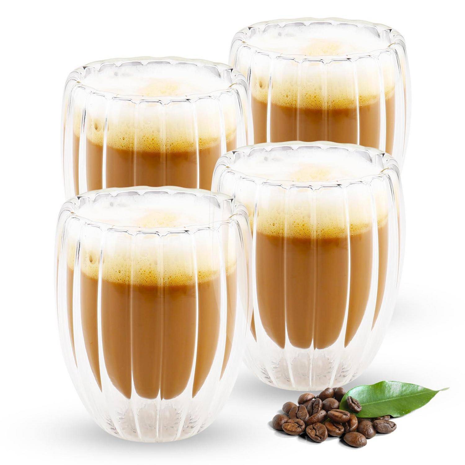 Vier klare doppelwandige Gläser mit geschichtetem Kaffee, oben mit Schaum gekrönt, mit Kaffeebohnen und einem grünen Blatt davor.