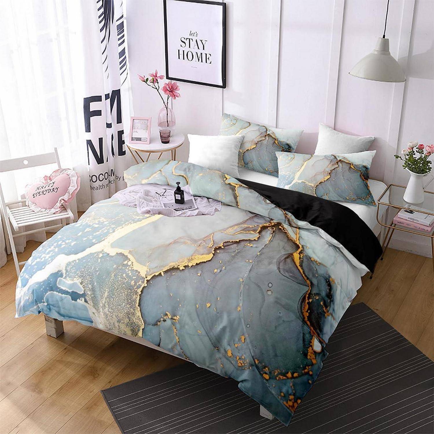 Ein ordentlich gemachtes Bett mit blau-goldenem Marmormuster-Bettwäsche, Kissen und einer schwarzen Wurfdecke in einem hellen Schlafzimmer mit Wandkunst und Nachttisch-Dekoration.