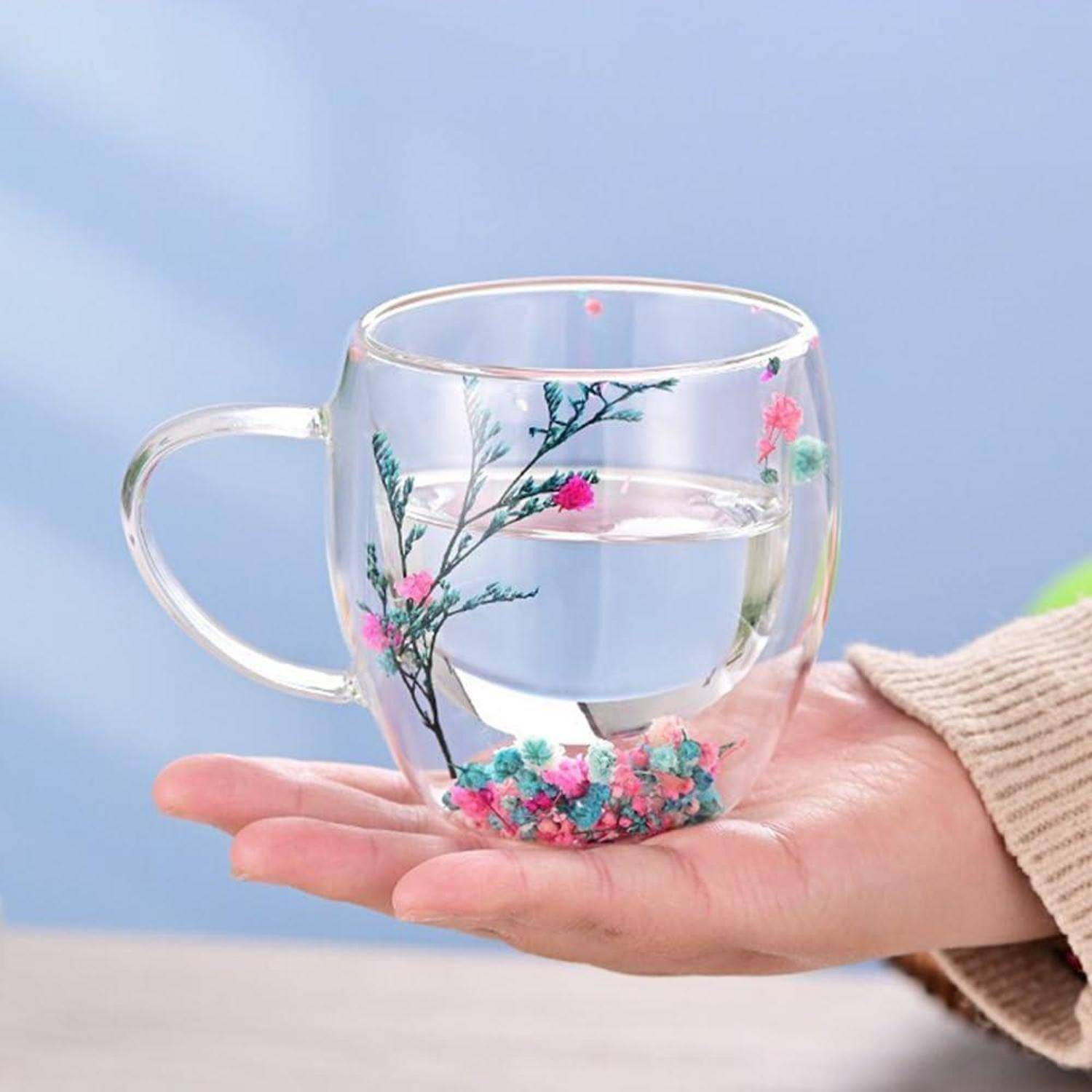 Hand, die einen klaren Glaskrug mit Wasser hält, verziert mit rosa Blumen und blauen Kieselsteinen.