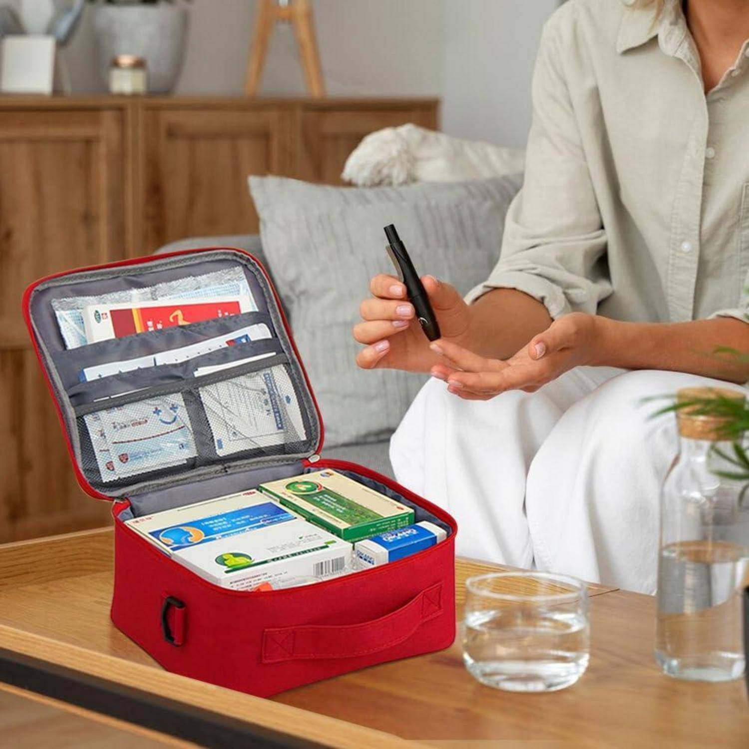 Person hält einen Stift neben einem offenen roten Erste-Hilfe-Kasten auf einem Couchtisch und zeigt organisierte Medizinboxen und Verbrauchsmaterialien in einem Wohnzimmer.