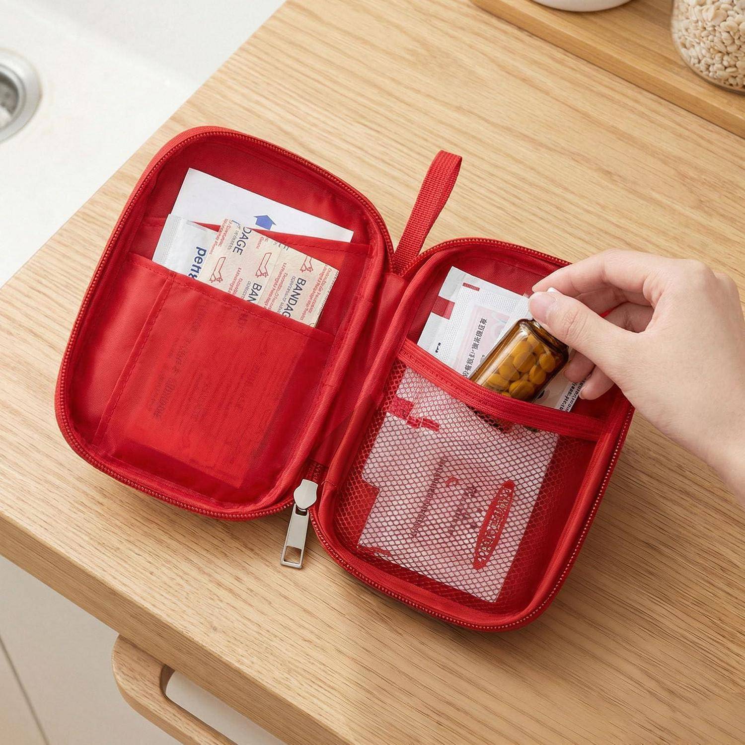 Hand legt eine Pillenflaschen in einen roten Reißverschluss-Organizer mit Netztaschen und Medikamentenpäckchen auf einem Holztisch.