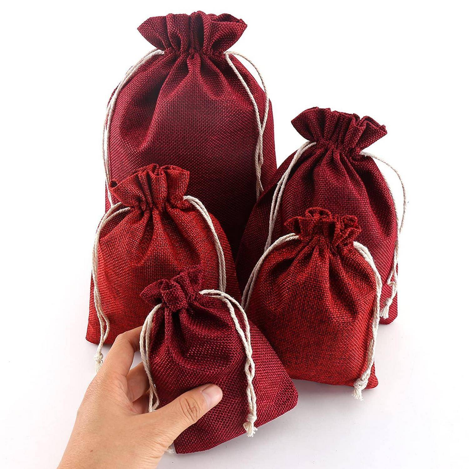 Fünf burgunderrote Geschenktaschen aus Stoff mit Kordelzug in verschiedenen Größen, wobei eine Hand die kleinste Tasche hält.
